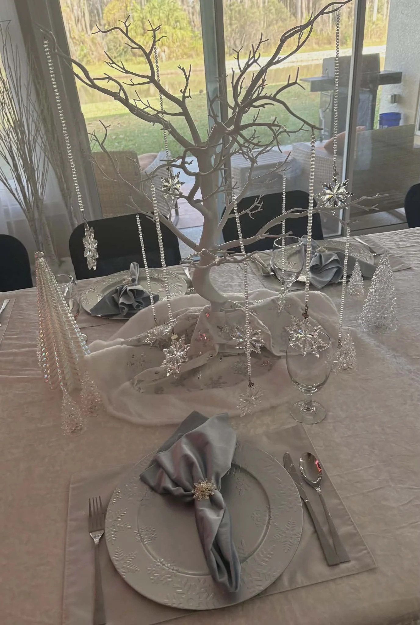 A decorated dining table featuring a centerpiece with a white tree adorned with hanging glass snowflakes, star, and bead ornaments in a bright room with large glass doors showing an outdoor grassy area.