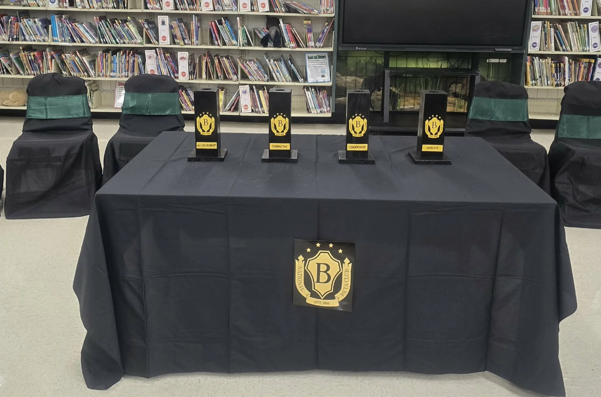 A table covered with a black cloth displaying four awards with the letter B, titles include Achievement, Character, Leadership, and Service, in a library with chairs covered in black slipcovers and bookshelves in the background.