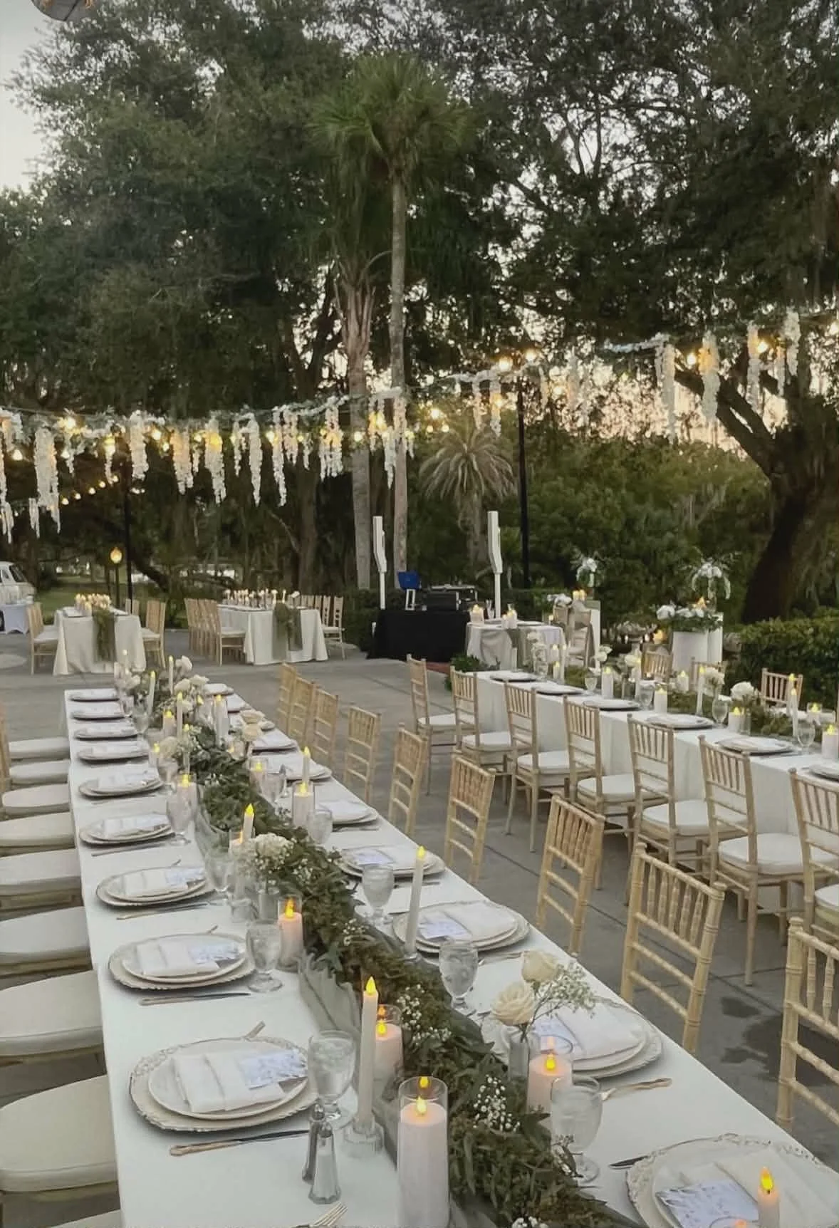Outdoor evening wedding reception setup with long dining tables adorned with candles, flowers, and white table settings under hanging string lights surrounded by trees.