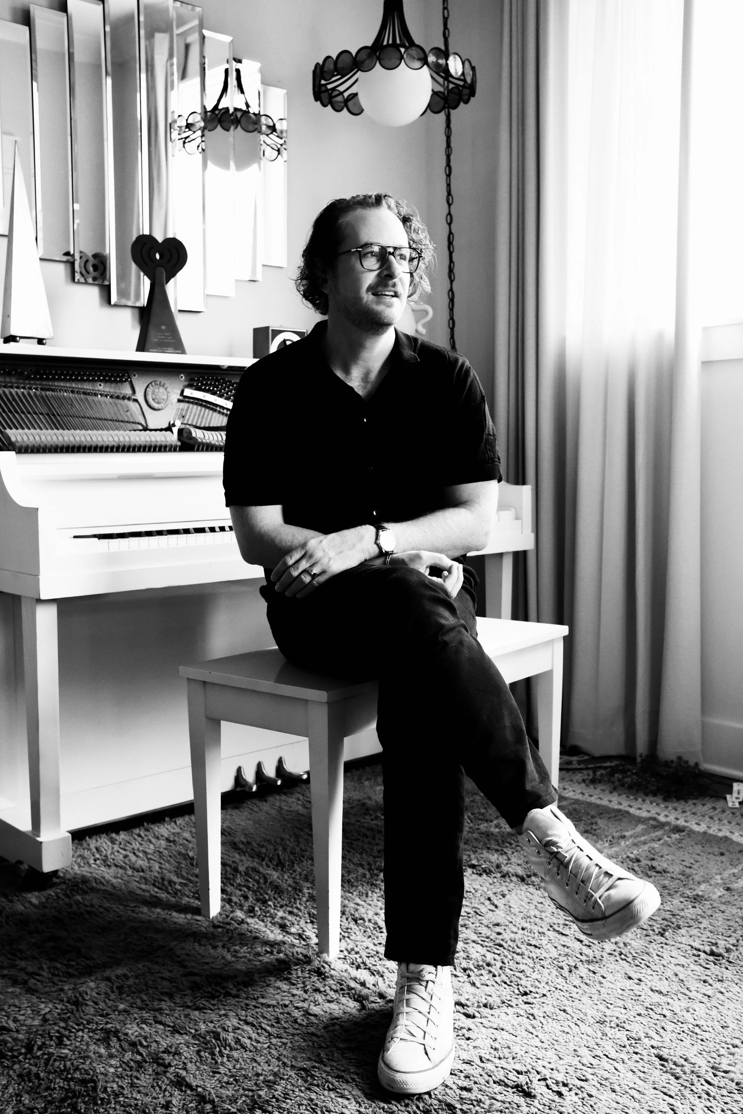 A man with glasses and curly hair sitting on a chair in a room with a white piano, decorative wall mirrors, and hanging lamps, looking off to the side.