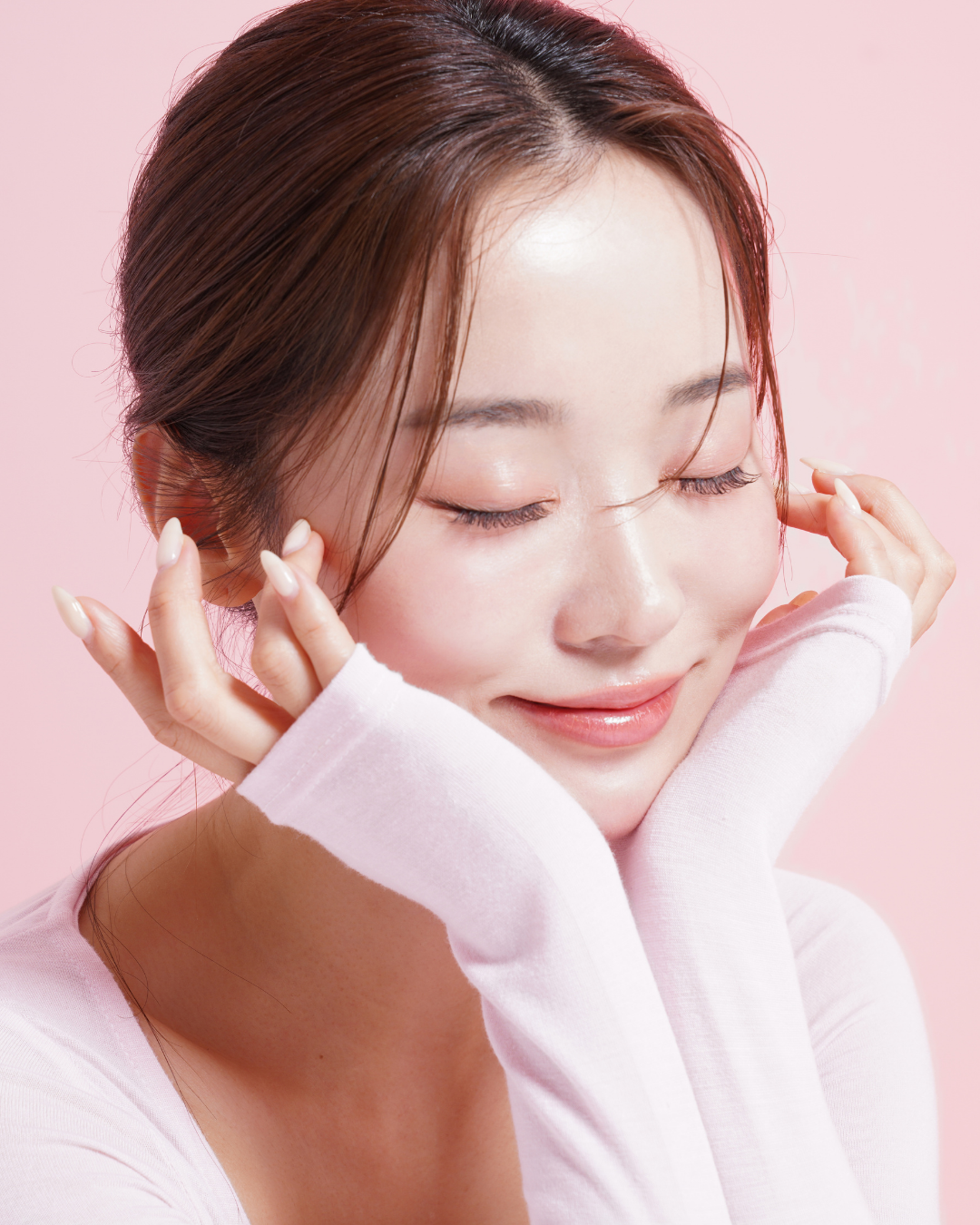 A young woman with closed eyes smiling, touching her face gently with both hands, wearing a light pink long-sleeve shirt, against a pink background.