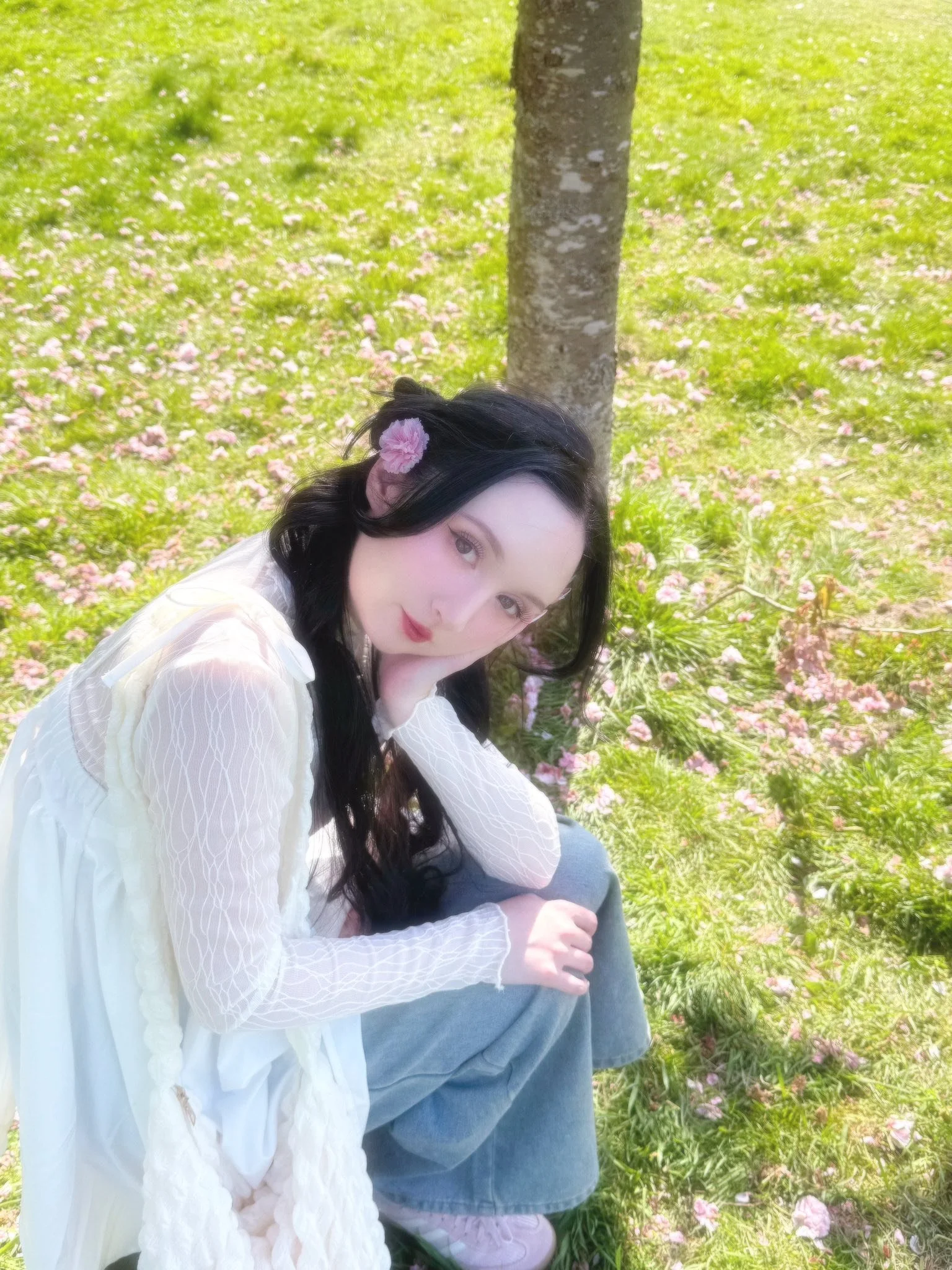 A woman with long black hair sitting on the grass next to a tree, wearing a white dress and a pink flower in her hair, surrounded by pink petals on the ground, in a park setting