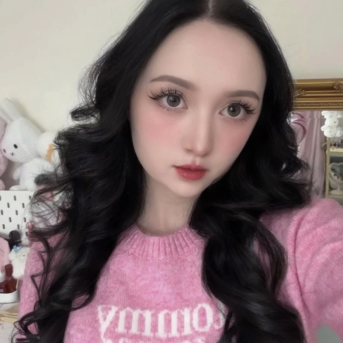 A young woman with long black wavy hair taking a selfie in a pink sweater with the word 'COOKIE' on it, standing in a room with plush toys and furniture in the background.