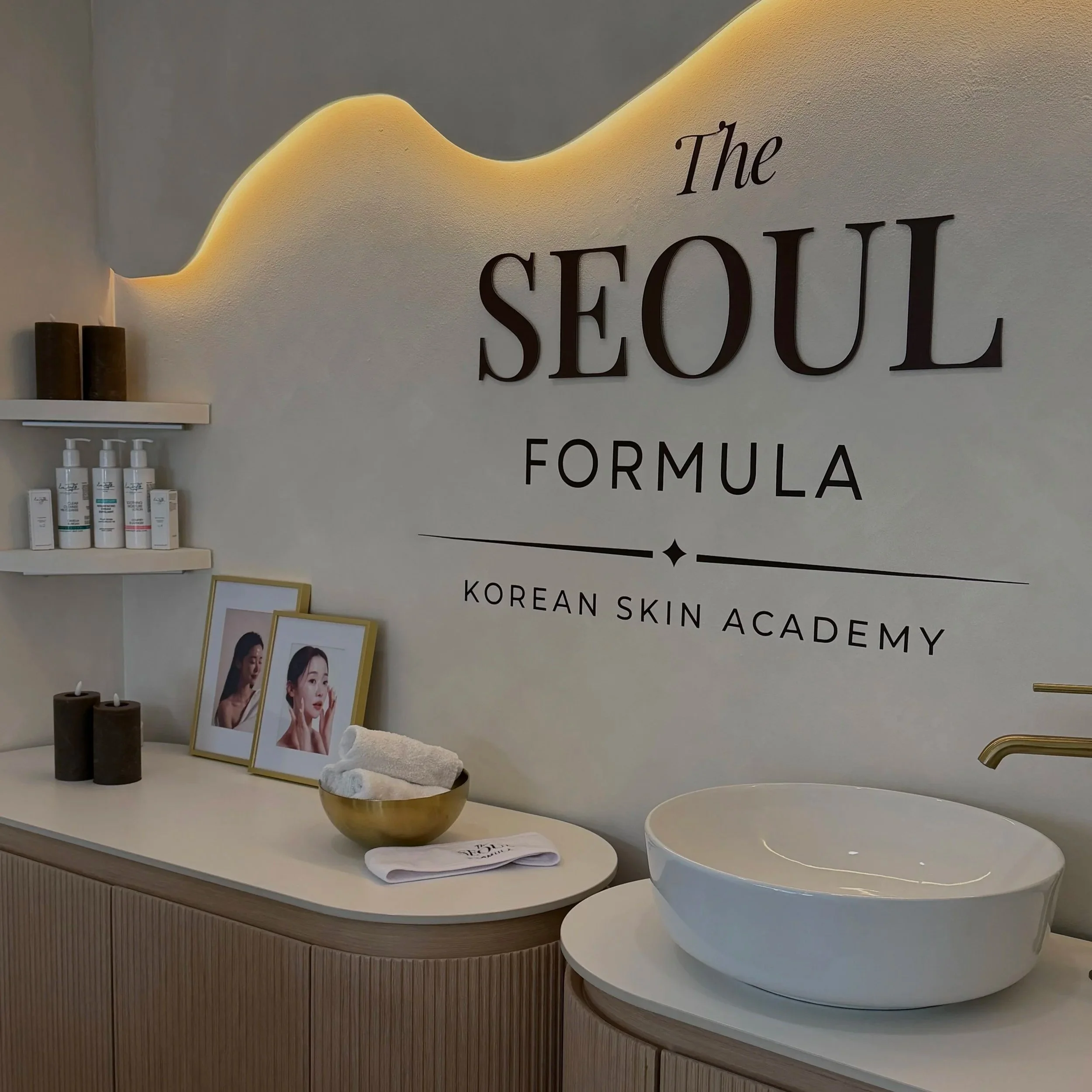 Interior of a skincare clinic with the sign 'The Seoul Formula Korean Skin Academy' on the wall, shelves with skincare products, framed photos, rolled towels, candles, and a modern sink.