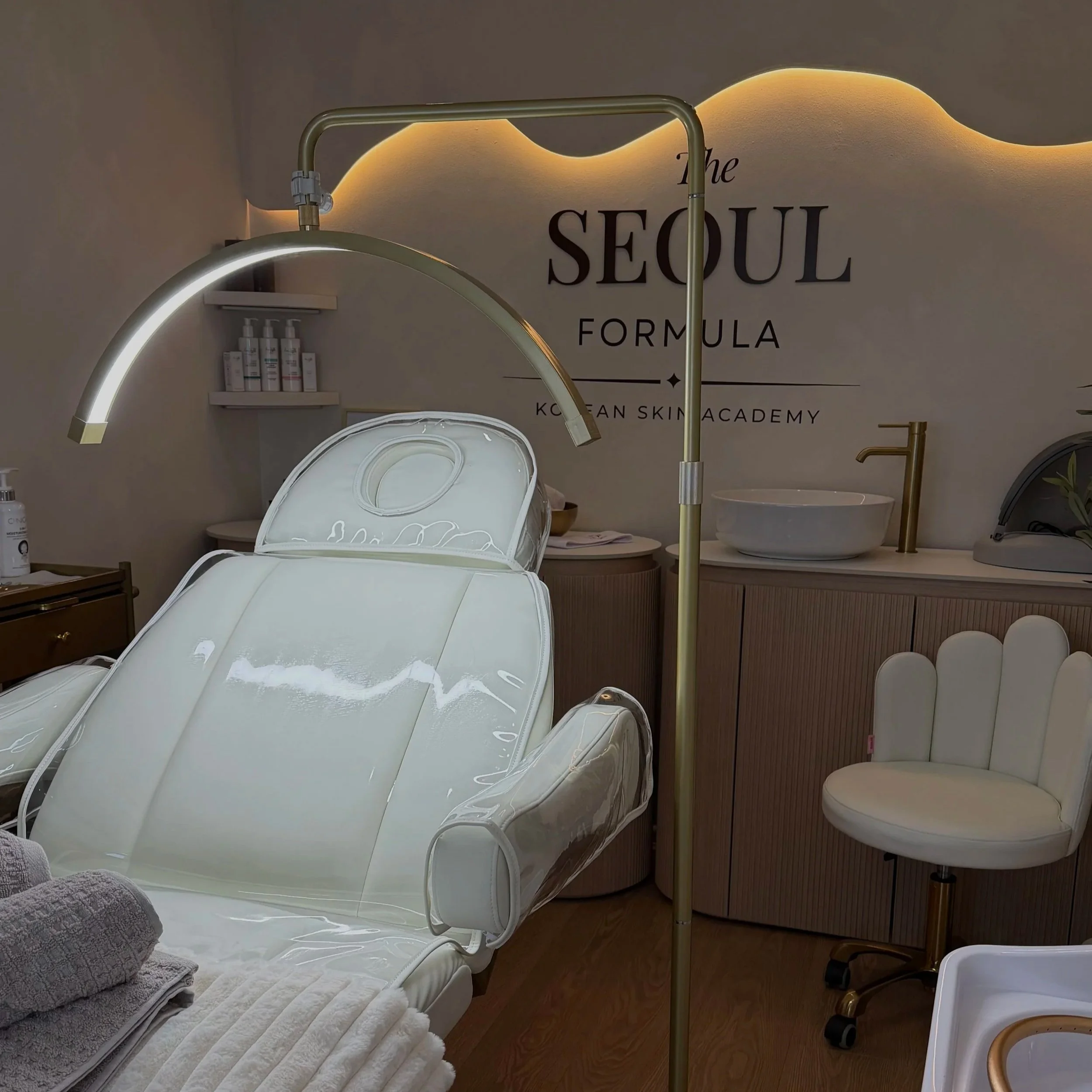 A treatment room at a Korean skincare clinic with a white reclining treatment chair, a gold and white medical lamp, a sign on the wall that says 'The Seoul Formula Korean Skin Academy,' a white sink with a golden faucet, and a white fitted chair.