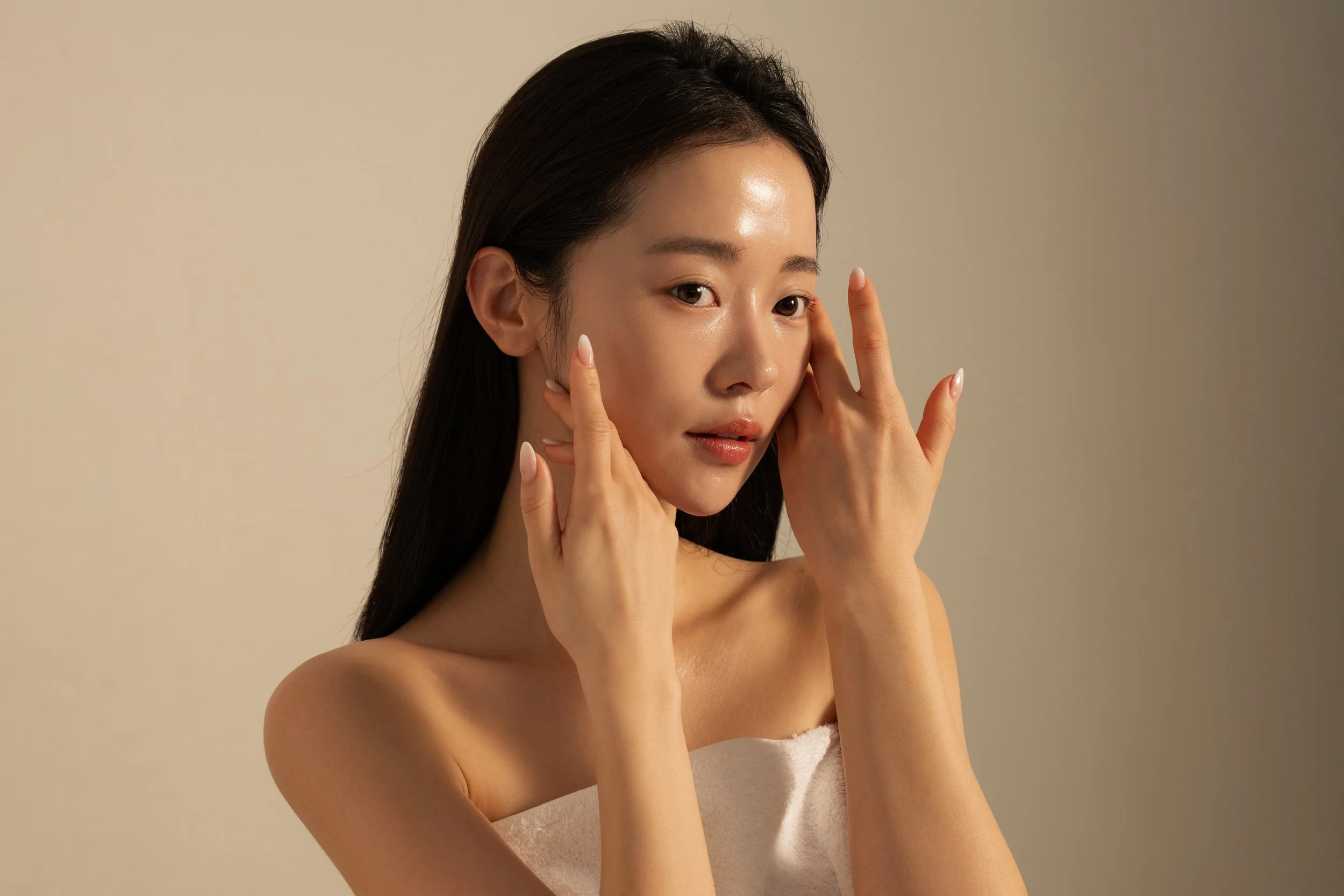 A young woman with long dark hair wrapped in a white towel posing with her hands near her face against a plain beige background.