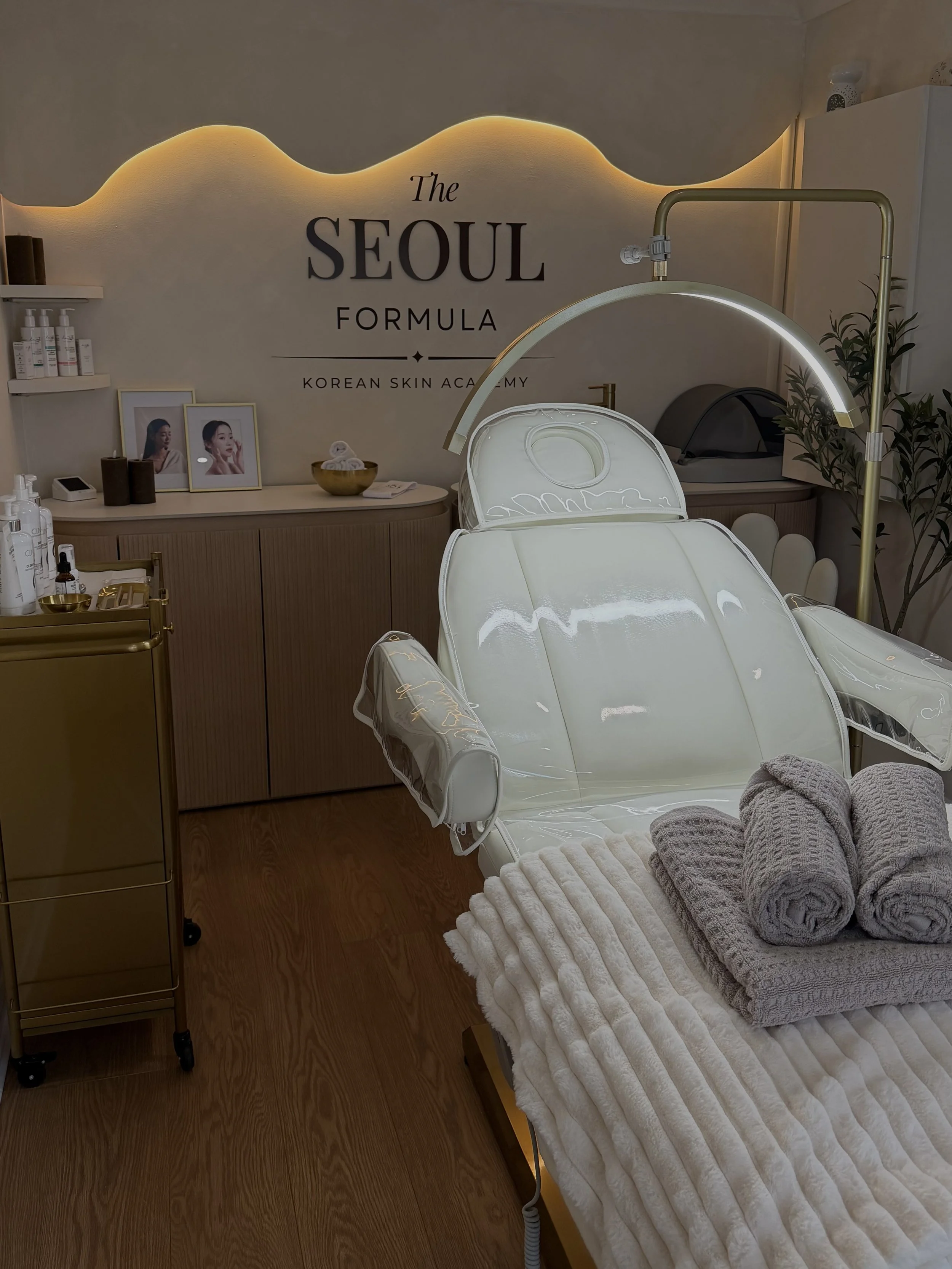 A massage or facial treatment room with a white treatment chair, rolled towels, and skincare products. The background features a wall with a sign that reads 'The Seoul Formula, Korean Skin Academy', and soft lighting.