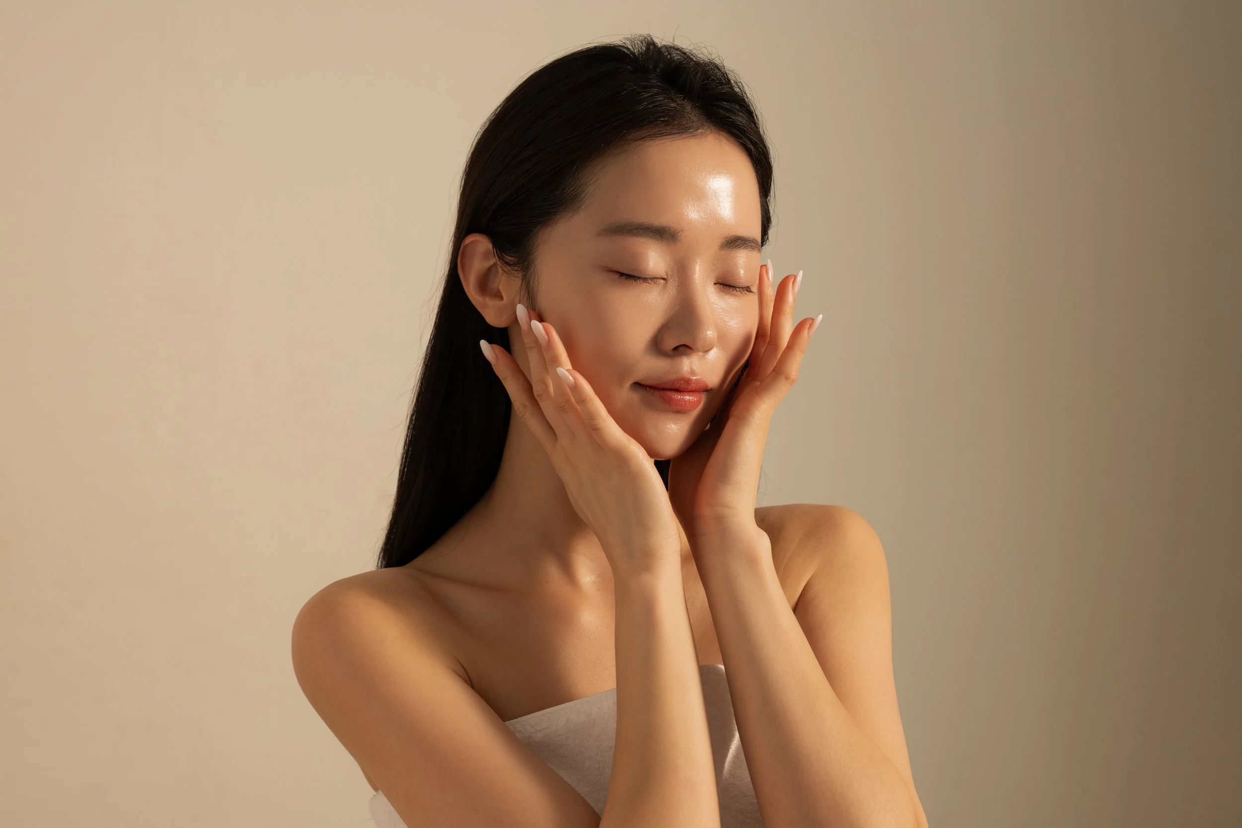 A woman with long dark hair gently touching her face with both hands, eyes closed, and a serene smile, against a neutral background.