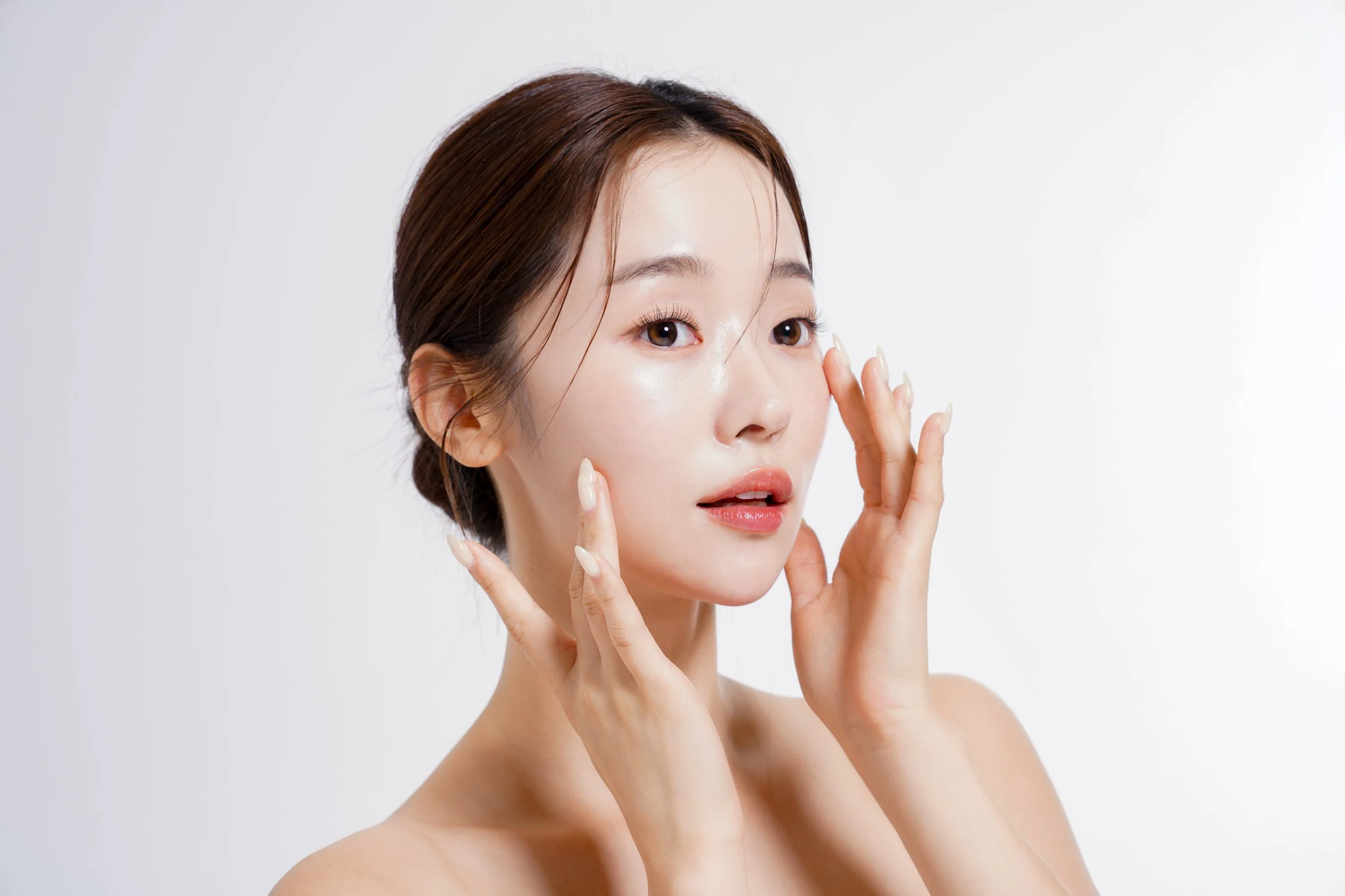 A woman with clear skin and brown hair styled in a bun, gently touching her face with her hands, against a plain white background.