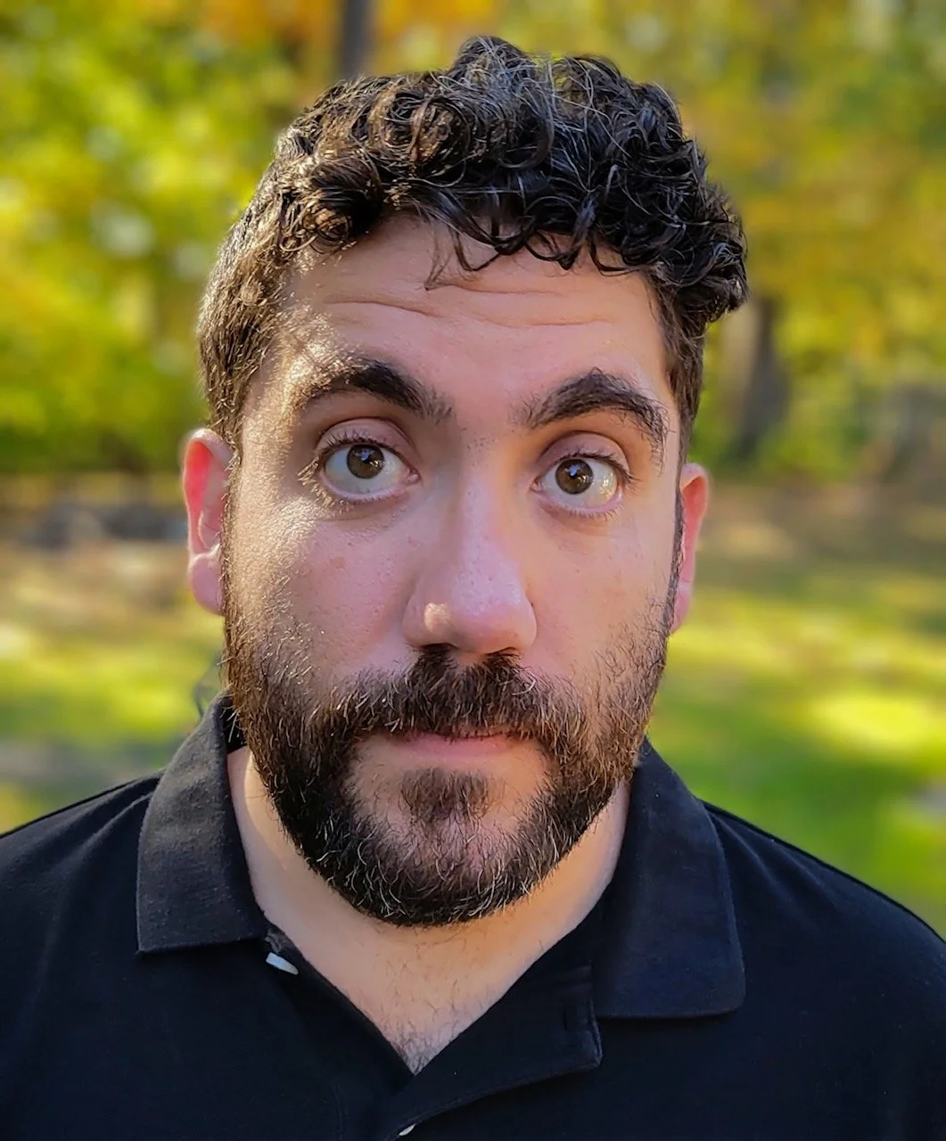 Close-up of a man with dark curly hair, brown eyes, and a beard, wearing a black polo shirt, outdoors with blurred green and yellow foliage in the background.