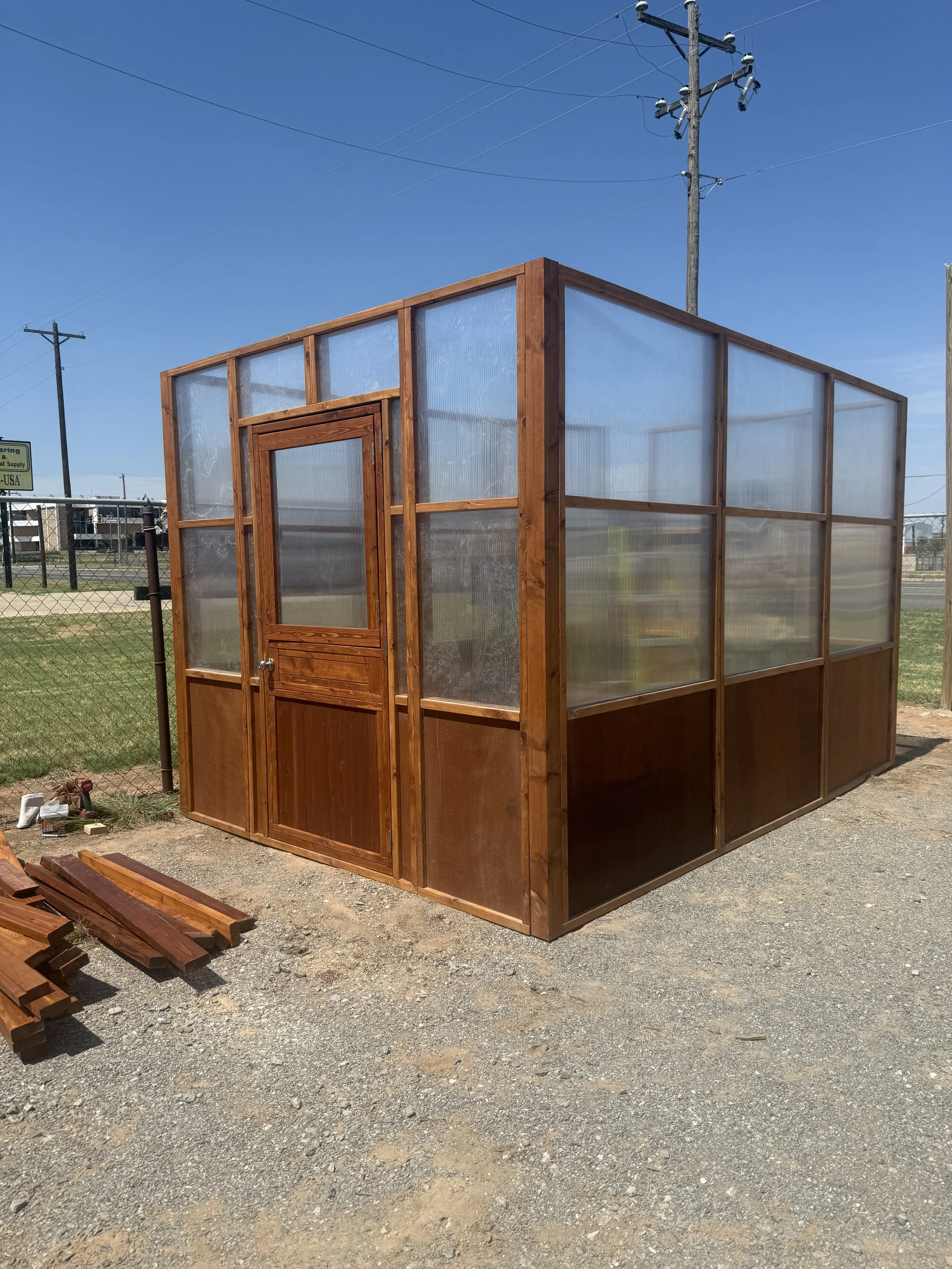 Stained cedar greenhouse on a gravel lot with stacked lumber nearby