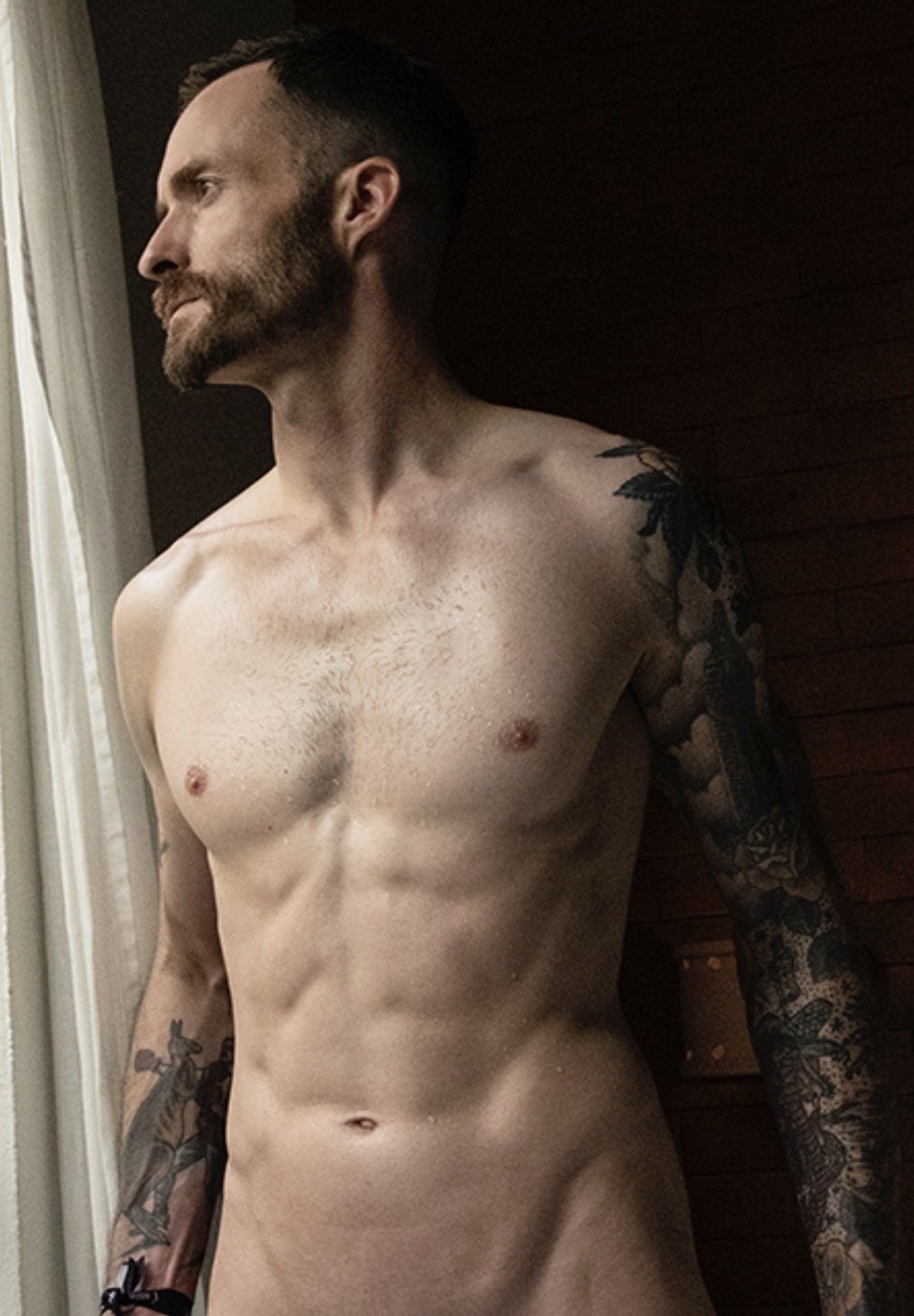 A shirtless man with tattoos on his left arm and right shoulder, looking to his left, standing indoors near a window with a dark wooden wall behind him.