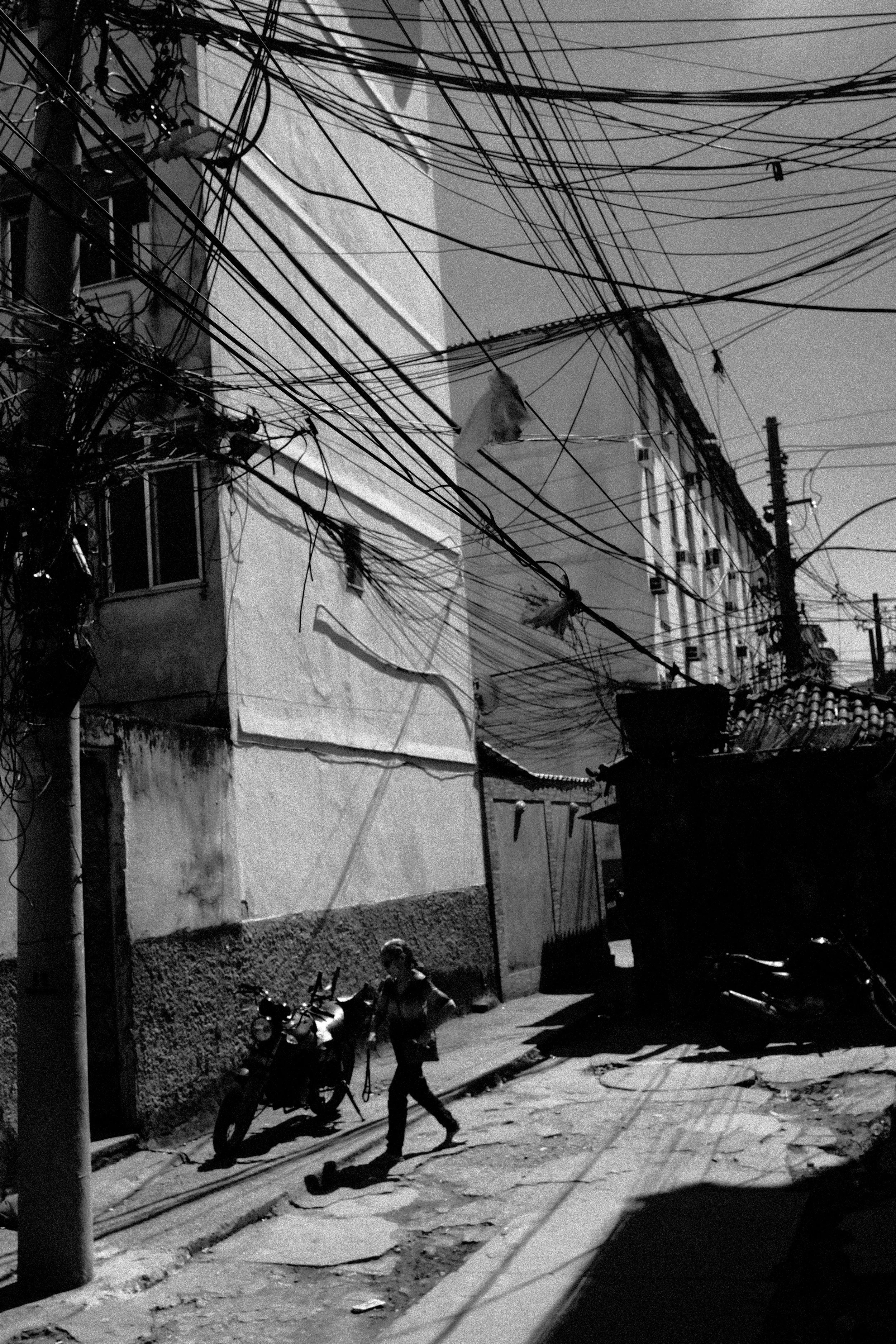 A black and white photo of a street scene with a building, tangled overhead wires, a man walking, and parked motorbikes.