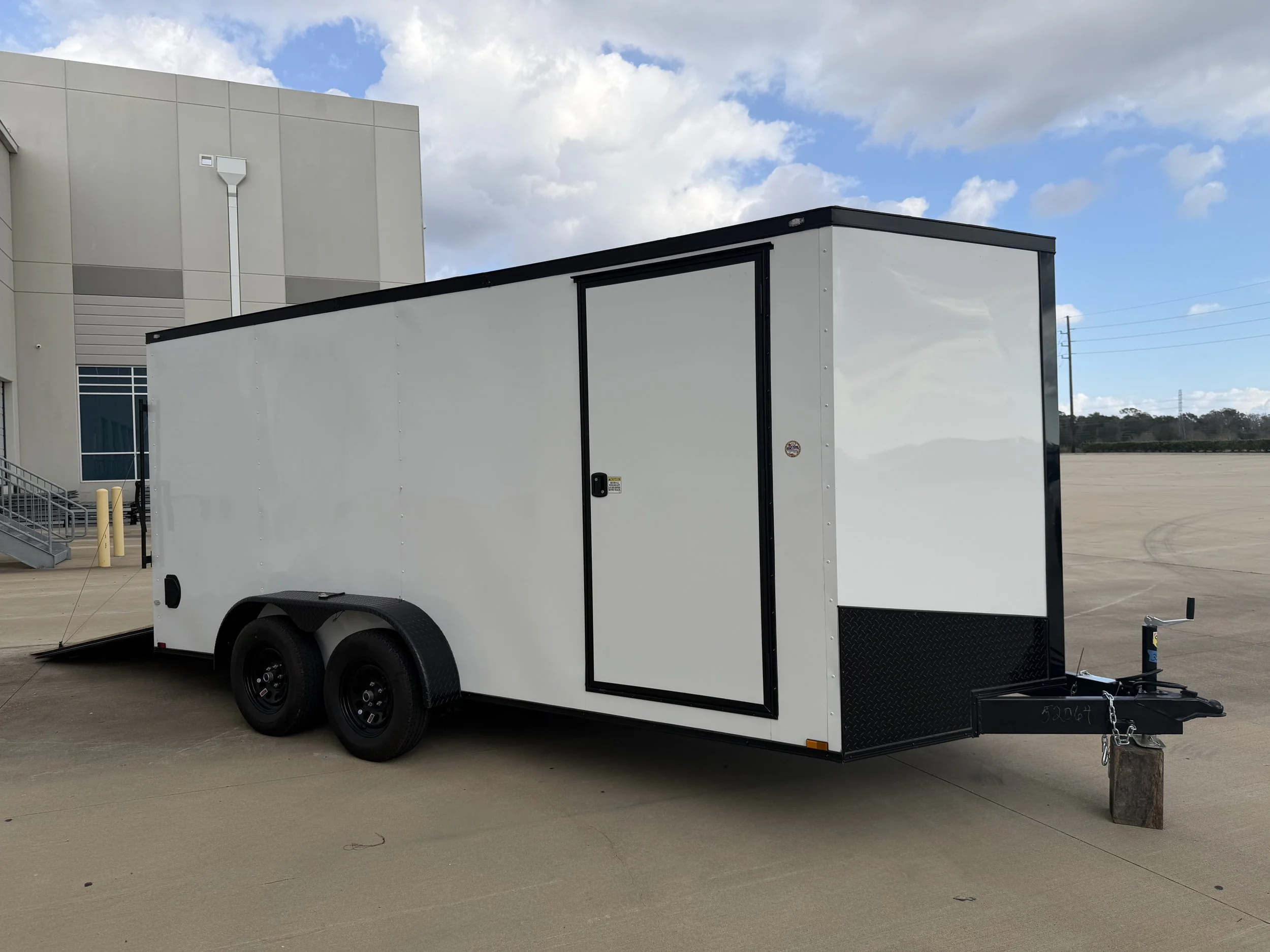 A white enclosed utility trailer with two black wheels parked on a concrete surface outside a building under a partly cloudy sky.