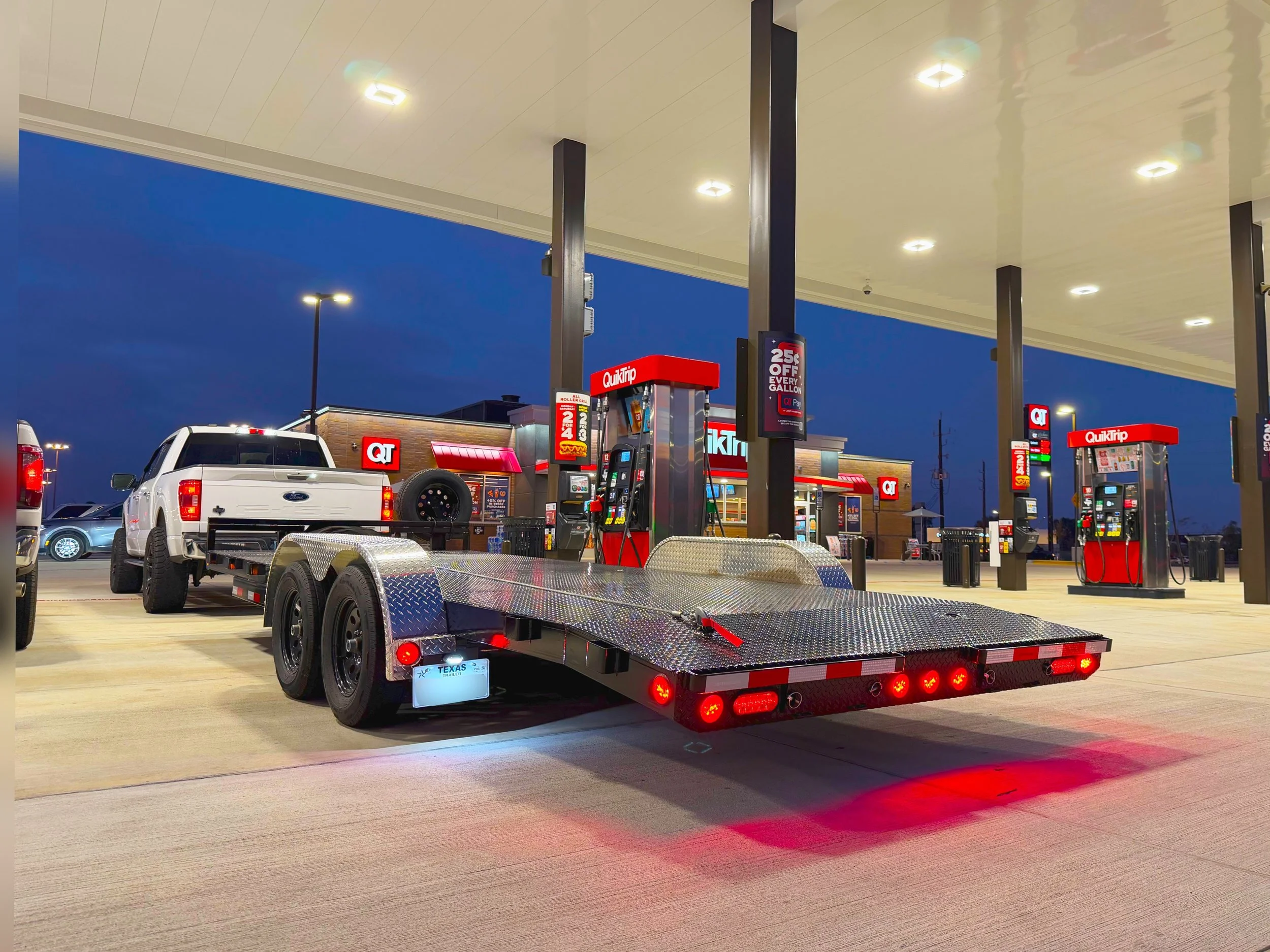Empty gas station at dusk with a flatbed trailer attached to a white pickup truck in the foreground, multiple fuel pumps, and a quick trip convenience store in the background.