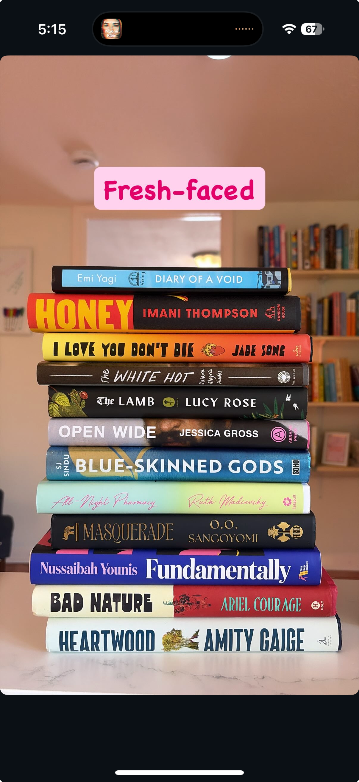 Stack of twelve books with colorful covers, titled from top to bottom: Diary of a Void, Honey, I Love You Don't Die, The White Hot, The Lamb, Open Wide, Blue-Skinned Gods, All-Night Pharmacy, Masquerade, Fundamentally, Bad Nature, Heartwood, Amity Gauge. The background shows a room with bookshelves and a light pink wall.