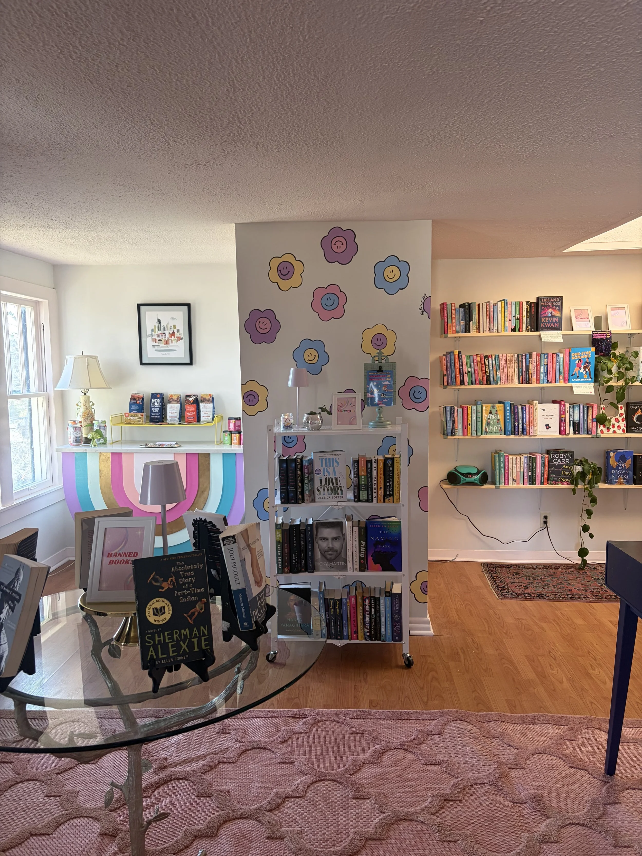 A cozy bookstore or library corner with a table displaying several books, a glass top on a carved wooden base, surrounded by a pink patterned rug. In the background, there are bookshelves filled with books and a corner with decorative wall art, a white side table, and a small yellow cart with additional books and decor items. The space is well-lit with natural light coming from a window.