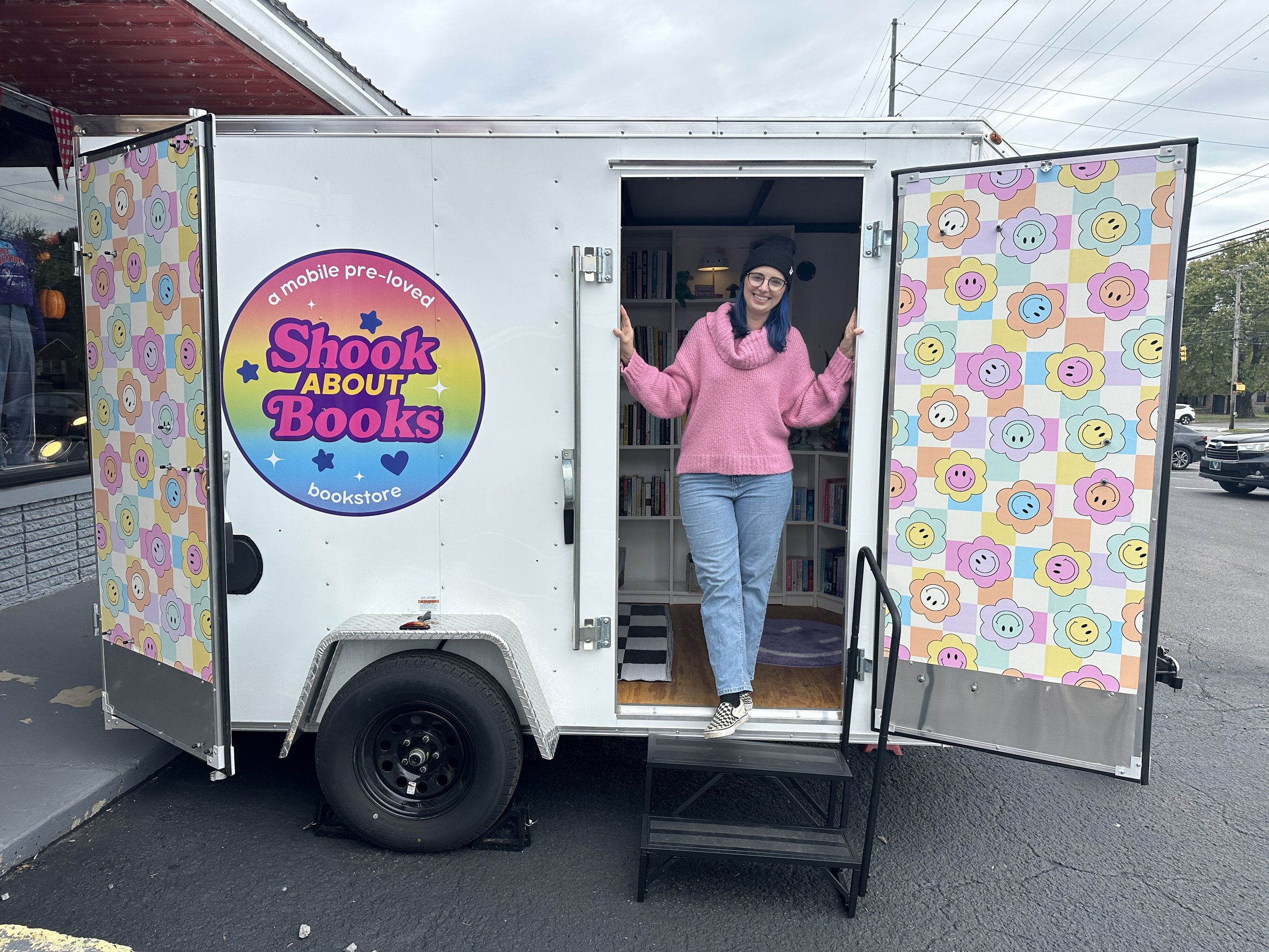 Zoë (owner) is standing in the doorway of a mobile bookstore called "Shook About Books" with colorful flower-patterned curtains on the open doors, smiling, wearing a pink sweater, jeans, and a black beanie.