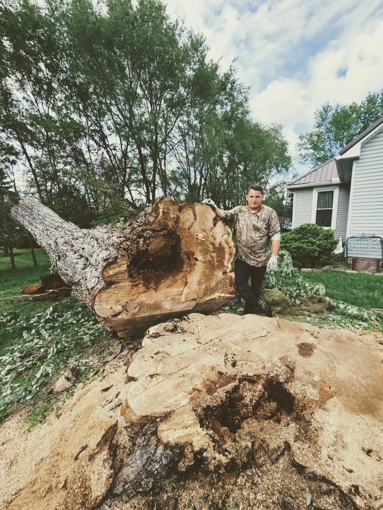 Man standing next to a large cut tree stump and a fallen tree in a residential yard with house and trees in the background.