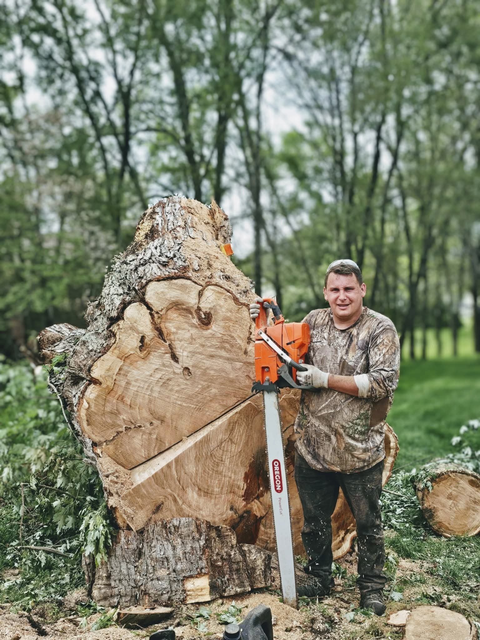 A man with camouflage clothing and gloves using a chainsaw to cut a large tree log outdoors, with a background of green trees and grass.