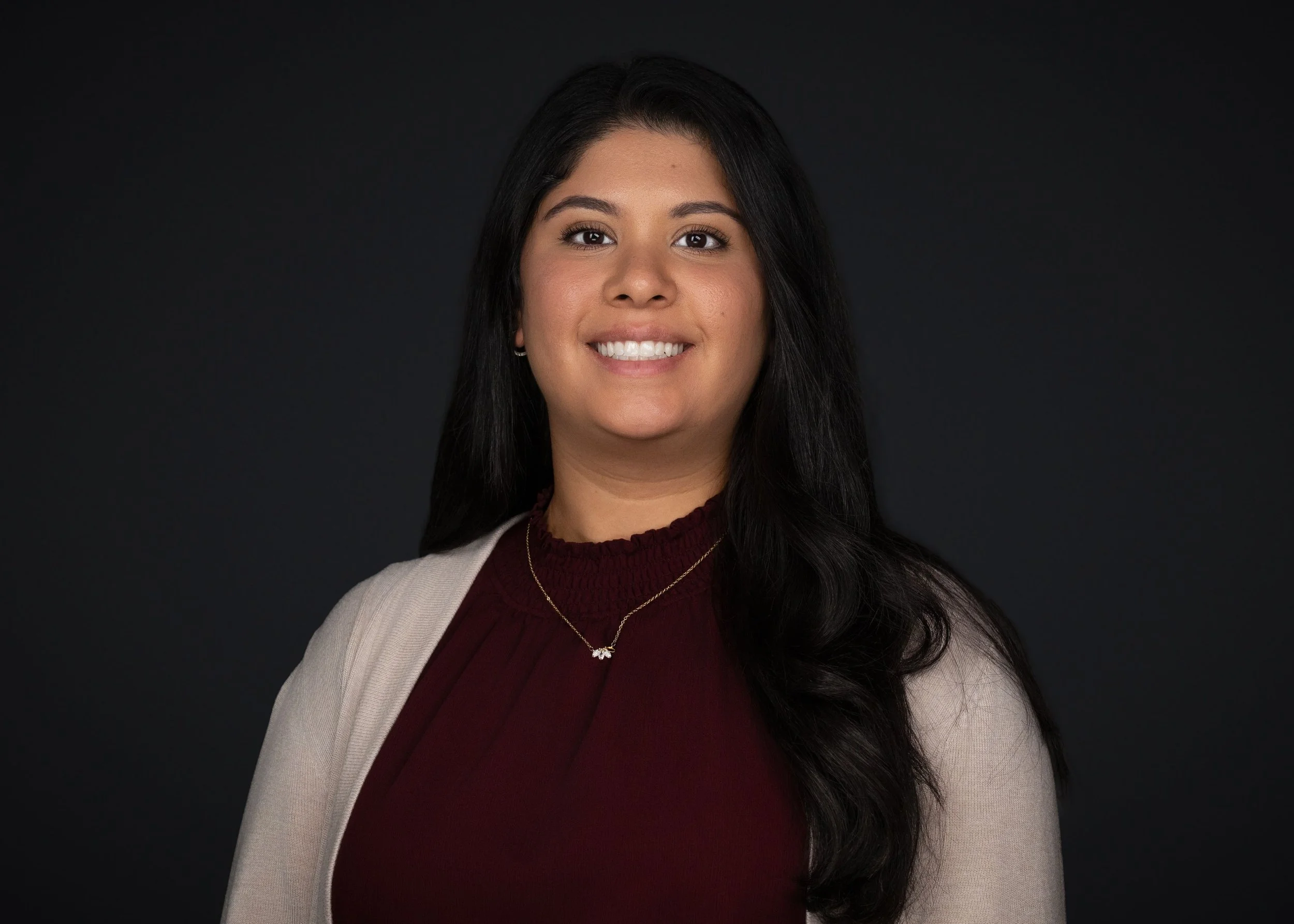 Portrait of a smiling woman with long black hair, wearing a beige blazer over a burgundy top, and a gold necklace, against a dark background.