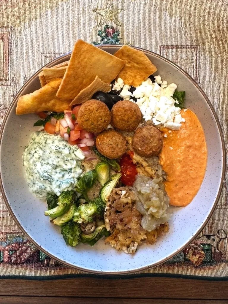 Plate of Mediterranean foods including falafel, pita bread, feta cheese, roasted vegetables, tzatziki sauce, and a red pepper spread, arranged on a speckled white dish.