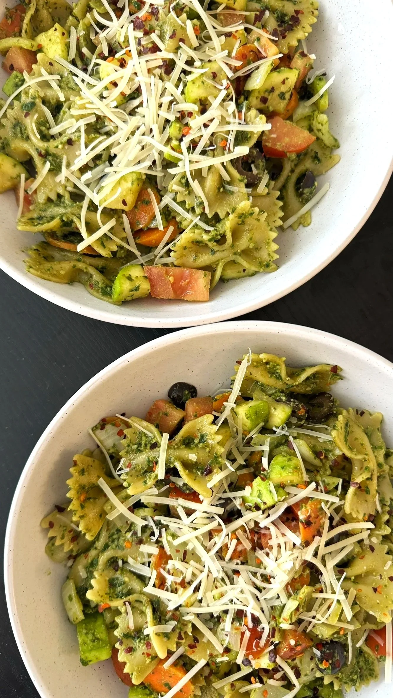 Two bowls of vegetable pasta salad topped with shredded cheese, containing farfalle and rotini pasta, cherry tomatoes, black olives, broccoli, and carrots.