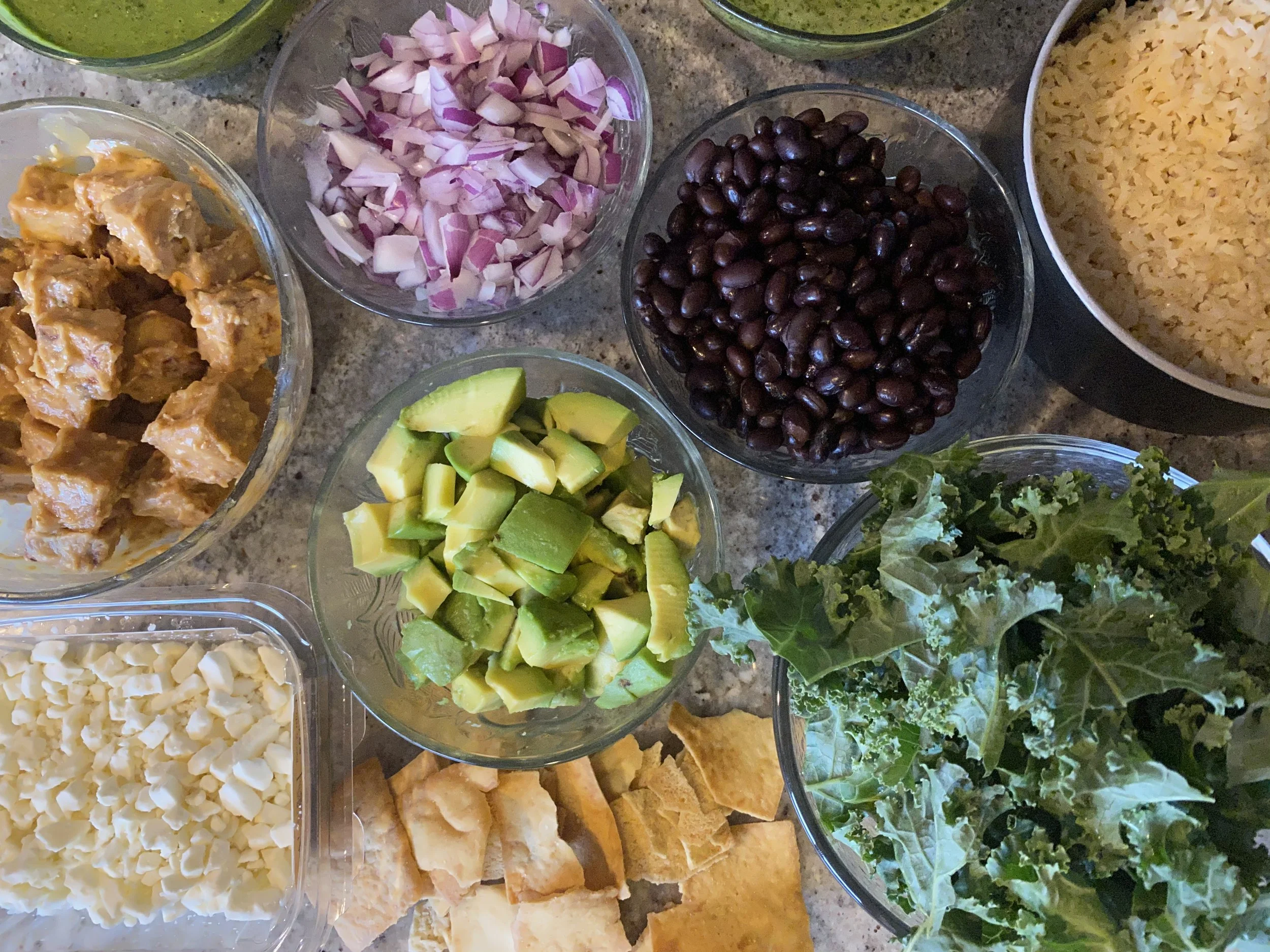 Bowls of ingredients for a salad or dish, including chopped red onion, black beans, shredded cheese, chopped avocado, chopped cooked chicken, chopped white cheese, tortilla chips, and leafy greens.