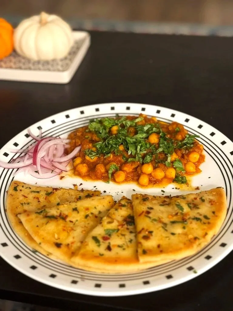 A plate with a chickpea curry, sliced naan bread garnished with herbs, and sliced red onions. In the background, small pumpkins are on a tray.