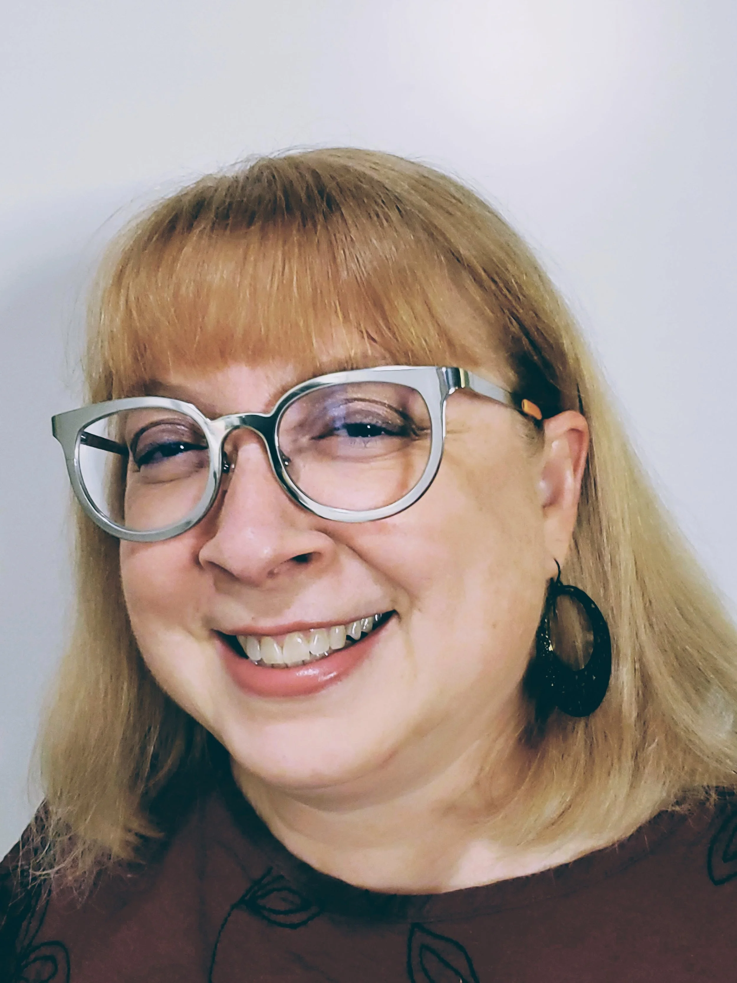 Close-up of a woman with blonde hair, wearing large glasses, smiling, and wearing a black earring.
