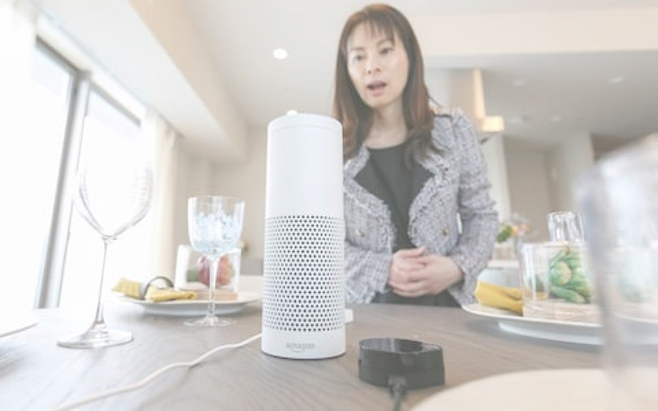 Woman speaking to a smart speaker on a dining table with glasses, plates, and see-through containers in the background.