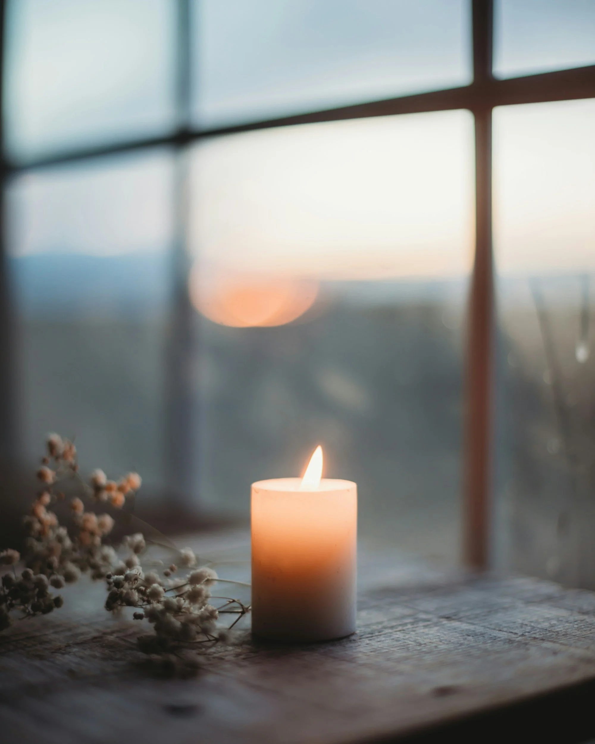 A lit candle on a wooden surface with a soft glow. Blurred flowers to the left and a window or mirror with divided panes in the background, showing a scenic view with bright sky reflecting the peaceful environment of Sagelight Counseling Studio.