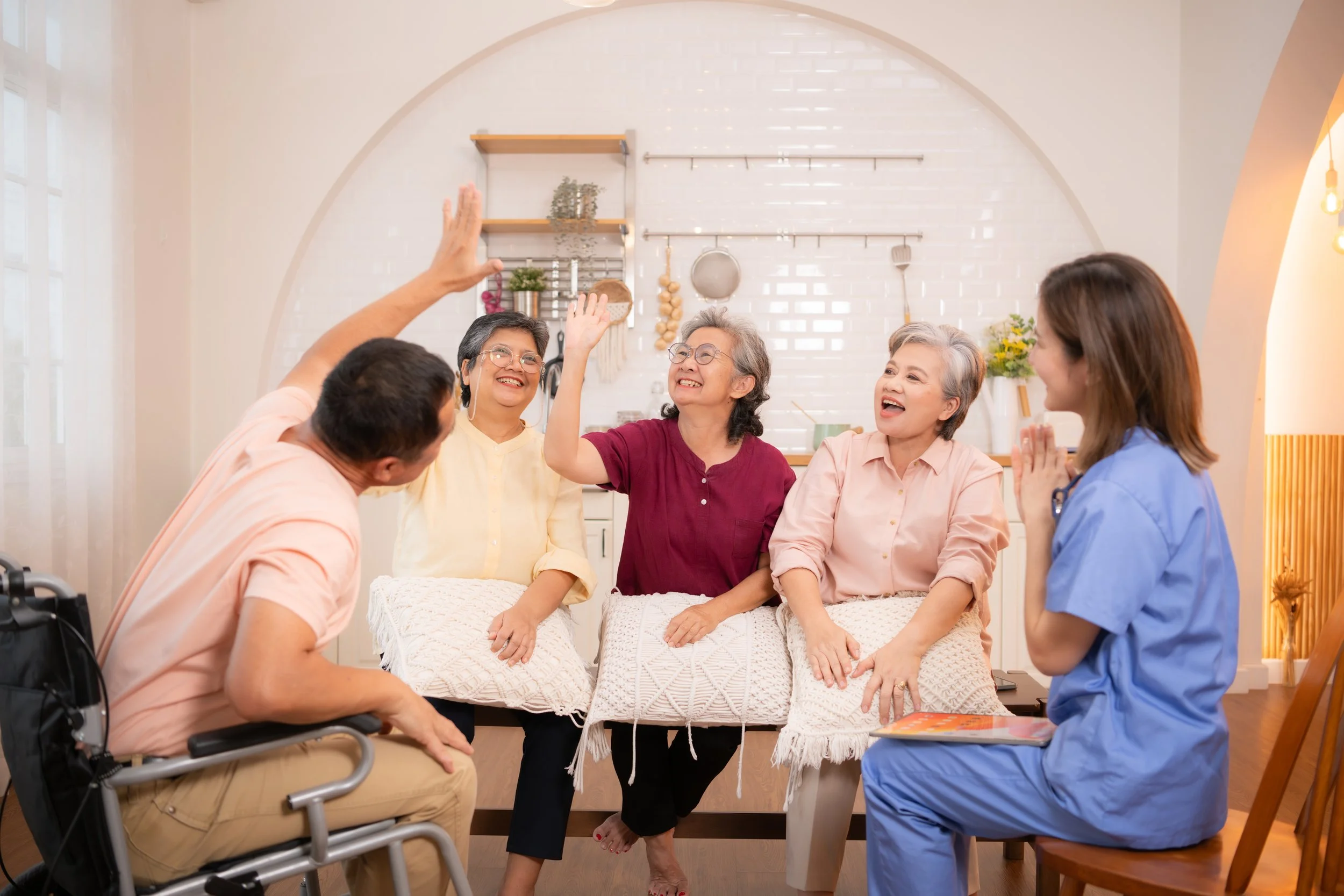 Group of elderly women and caregivers in a hospital or care home setting, engaging in a joyful high-five and conversation.