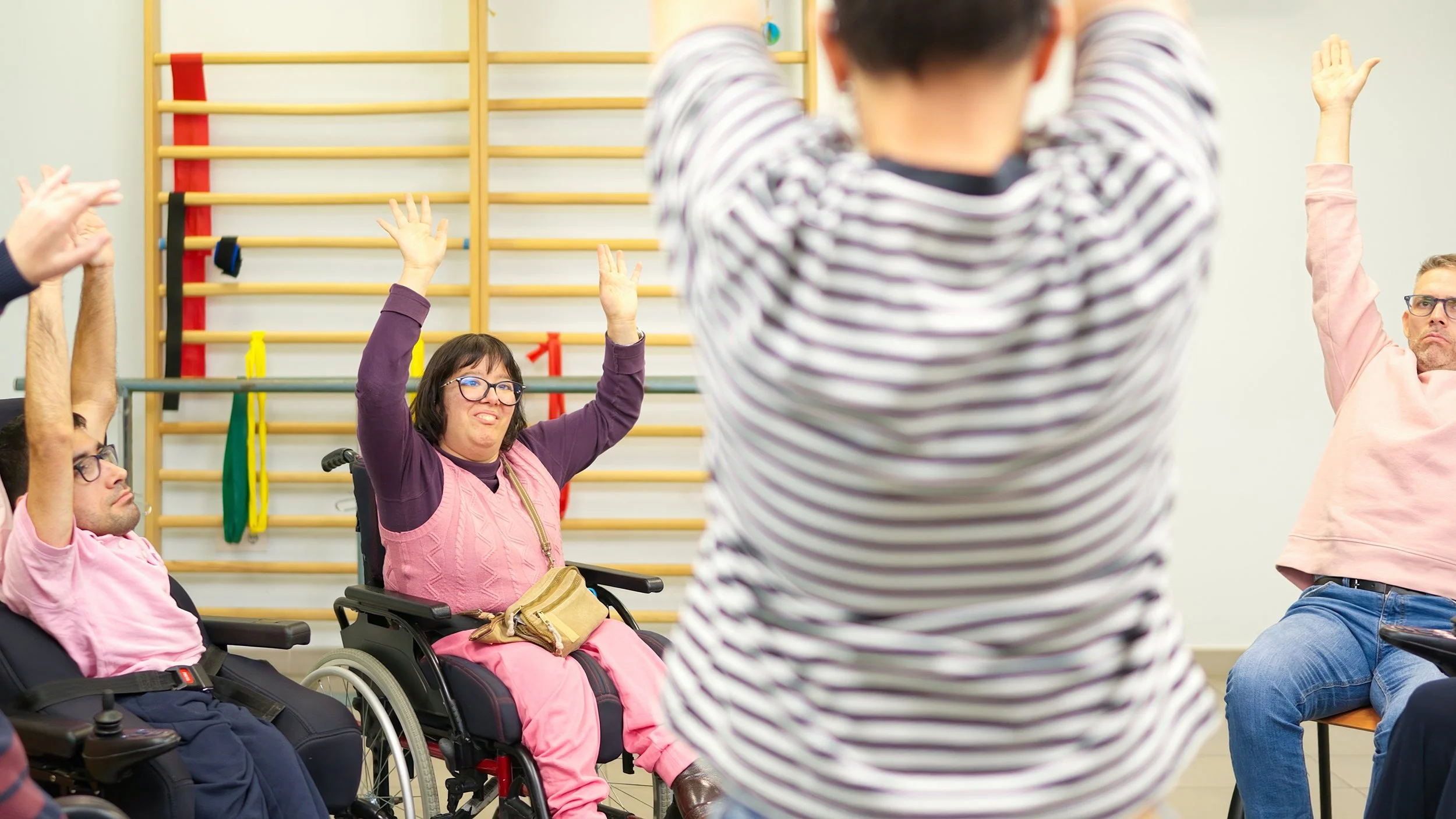Group of people, including individuals in wheelchairs, participating in a seated activity in a therapy or activity room. Some have their hands raised, possibly engaging in a group exercise or discussion.