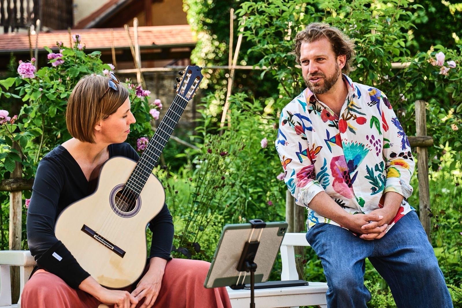 Zwei Menschen, eine Frau mit Akustikgitarre und ein Mann, unterhalten sich im Garten, im Hintergrund mit blühenden Rosen und Bäumen.
