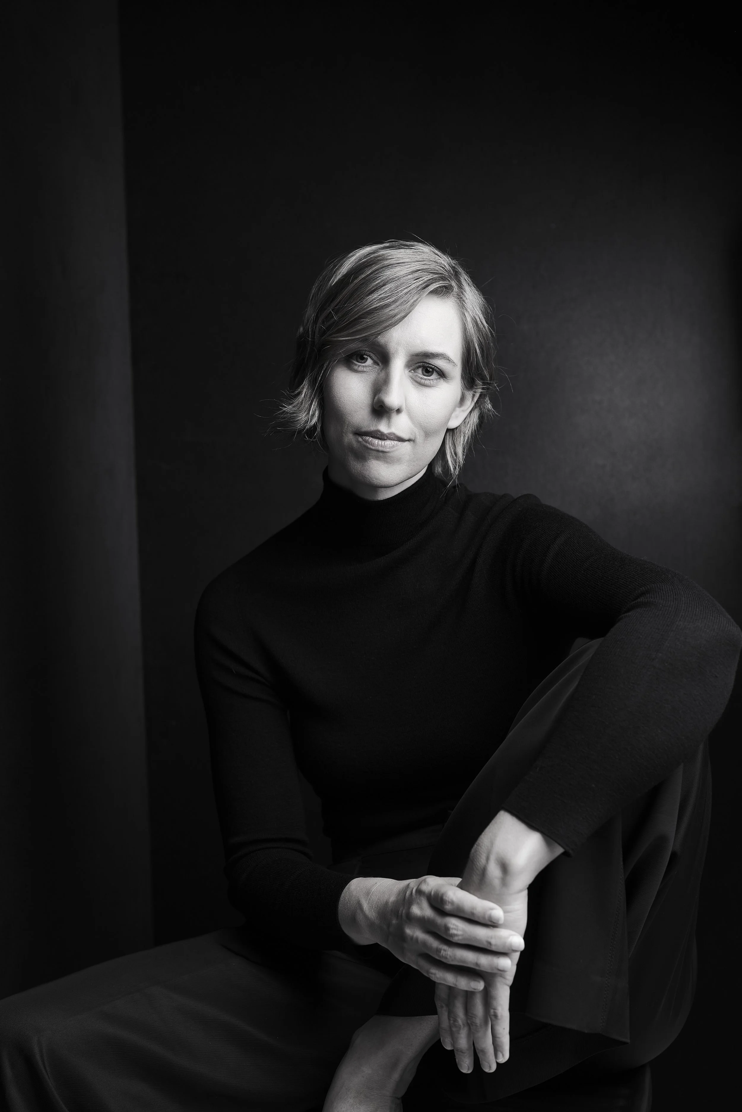 Schwarz-weiß Porträt einer jungen Frau mit kurzem Haar, die in schwarzer Kleidung auf einem dunklen Hintergrund sitzt.