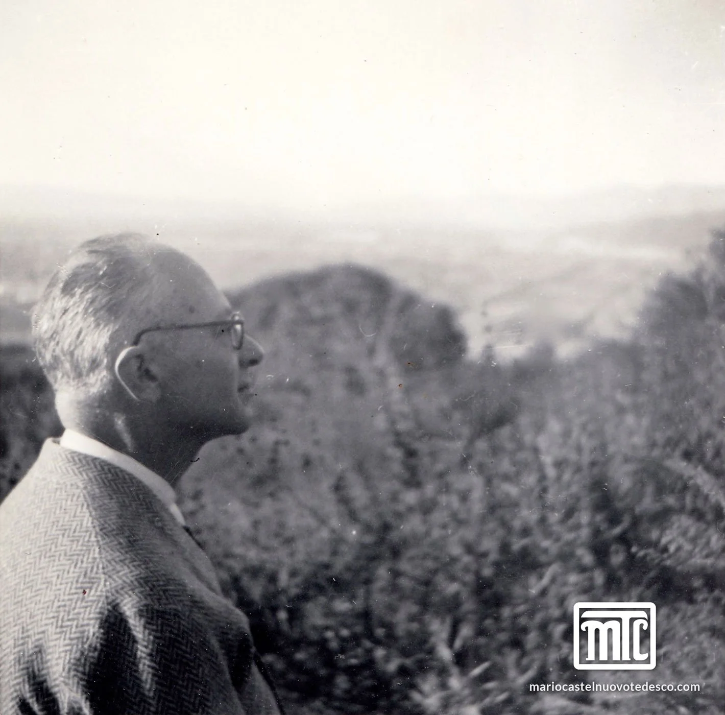 Schwarze und weiße Fotografie eines Mannes in Brille und Hemd, blickt nach rechts, im Hintergrund Bäume und Himmel.