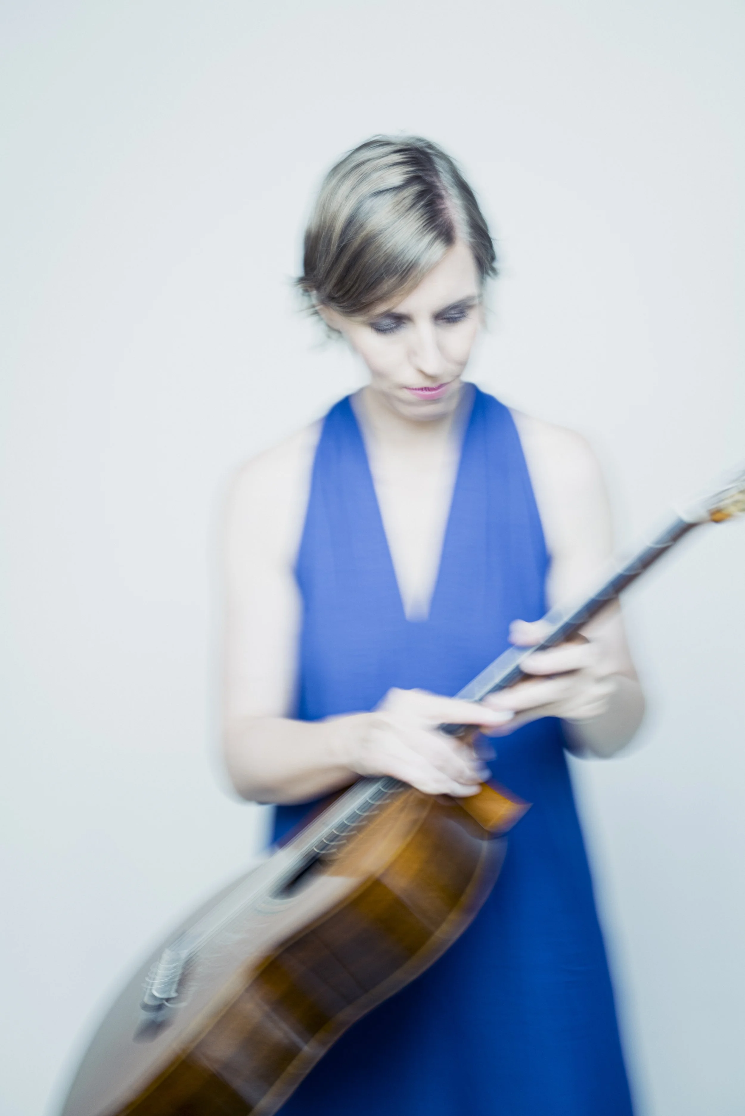 Frau in blauer Kleid mit E-Gitarre, Blick nach unten, kurzhaarige braune Haare, weiße Wand im Hintergrund