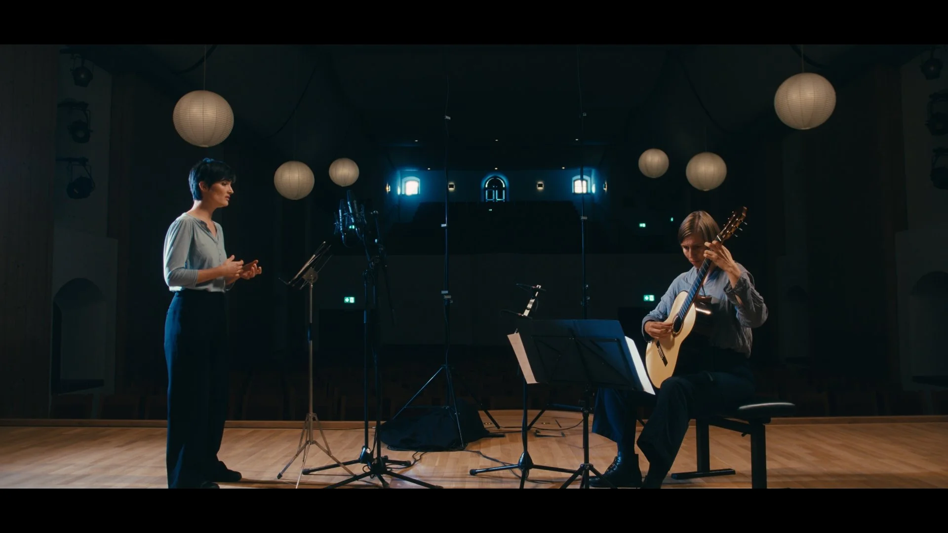 Zwei Frauen auf einer Bühne, eine singt und die andere spielt Gitarre bei einem Musik- oder Gesangsprojekt, mit Mikrofonen und Musiknotenständer, im Hintergrund dekorative Lampions.