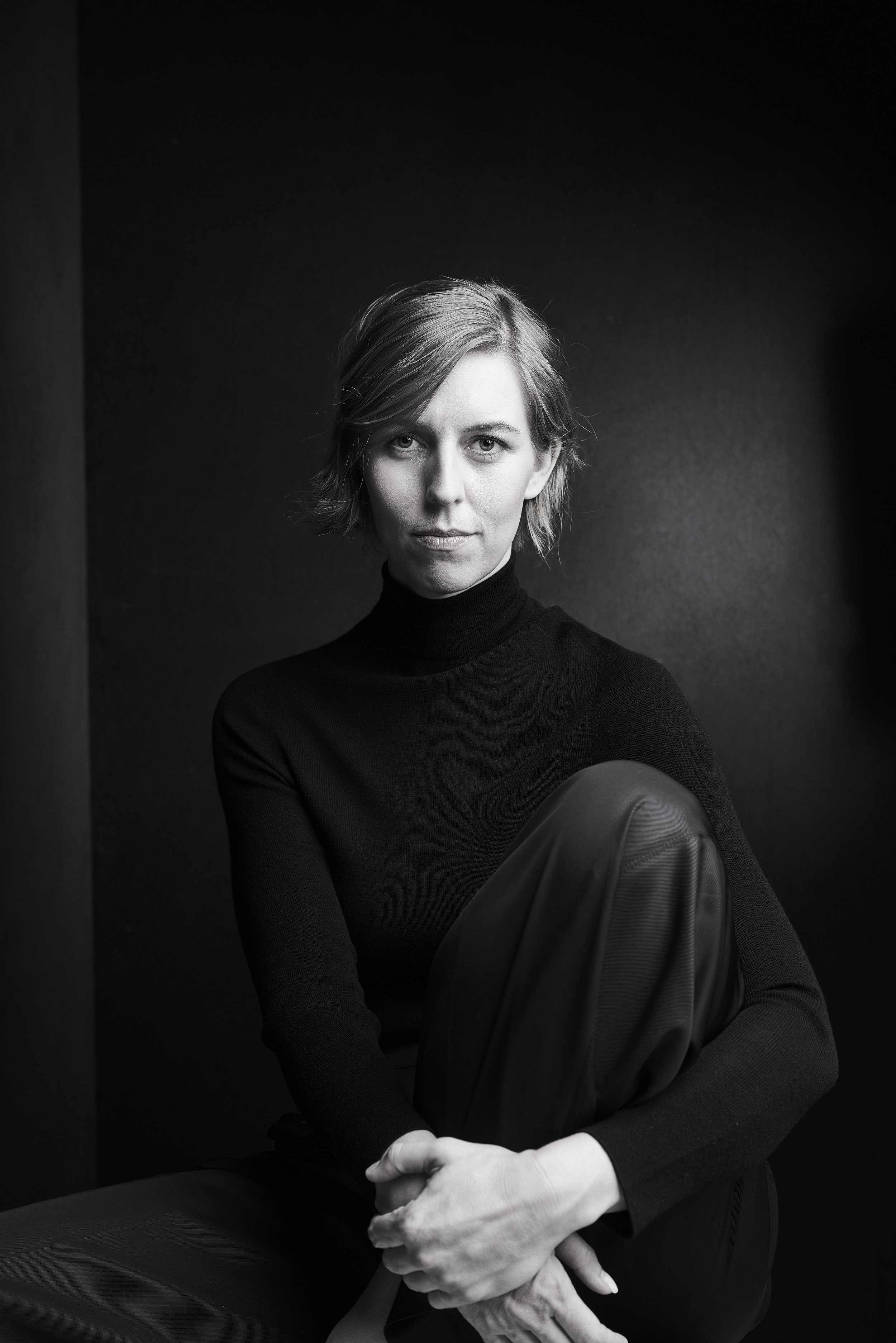 Ein Schwarz-Weiß-Foto einer Frau mit kurzem Haar, die in die Kamera schaut, schwarz Kleid und schwarze Hose trägt, sitzend mit einem Bein hochgezogen und den Arm um das Knie legend.