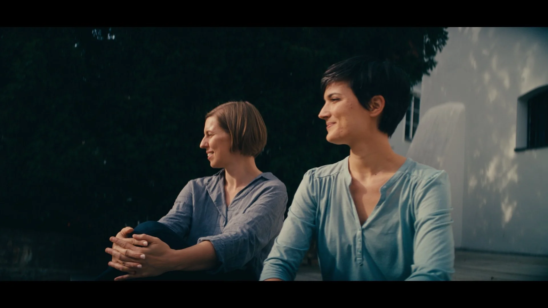 Zwei Frauen sitzen nebeneinander im Freien und unterhalten sich, eine lacht, während die andere lächelt, im Hintergrund ein Baum und ein weißes Gebäude.