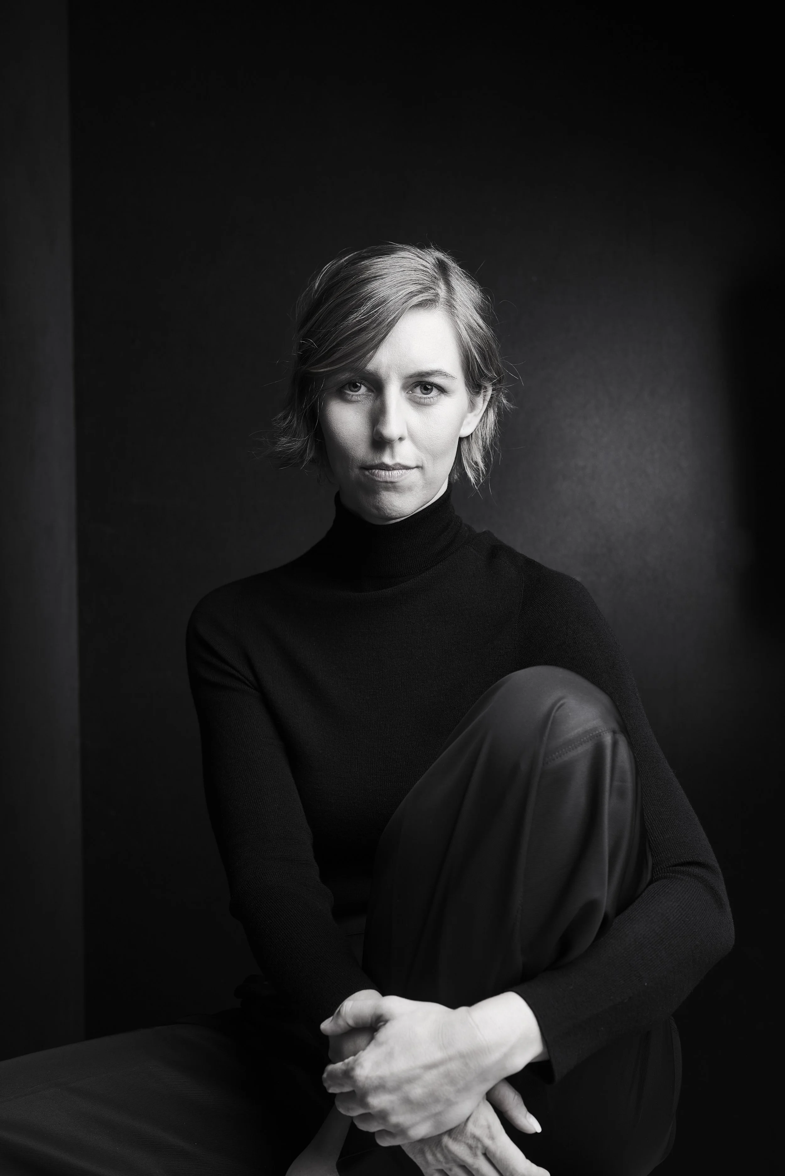 Schwarz-weiß Porträt einer jungen Frau mit Kurzhaarfrisur, die schwarze Kleidung trägt, vor dunklem Hintergrund, sitzend und den Blick direkt in die Kamera gerichtet.