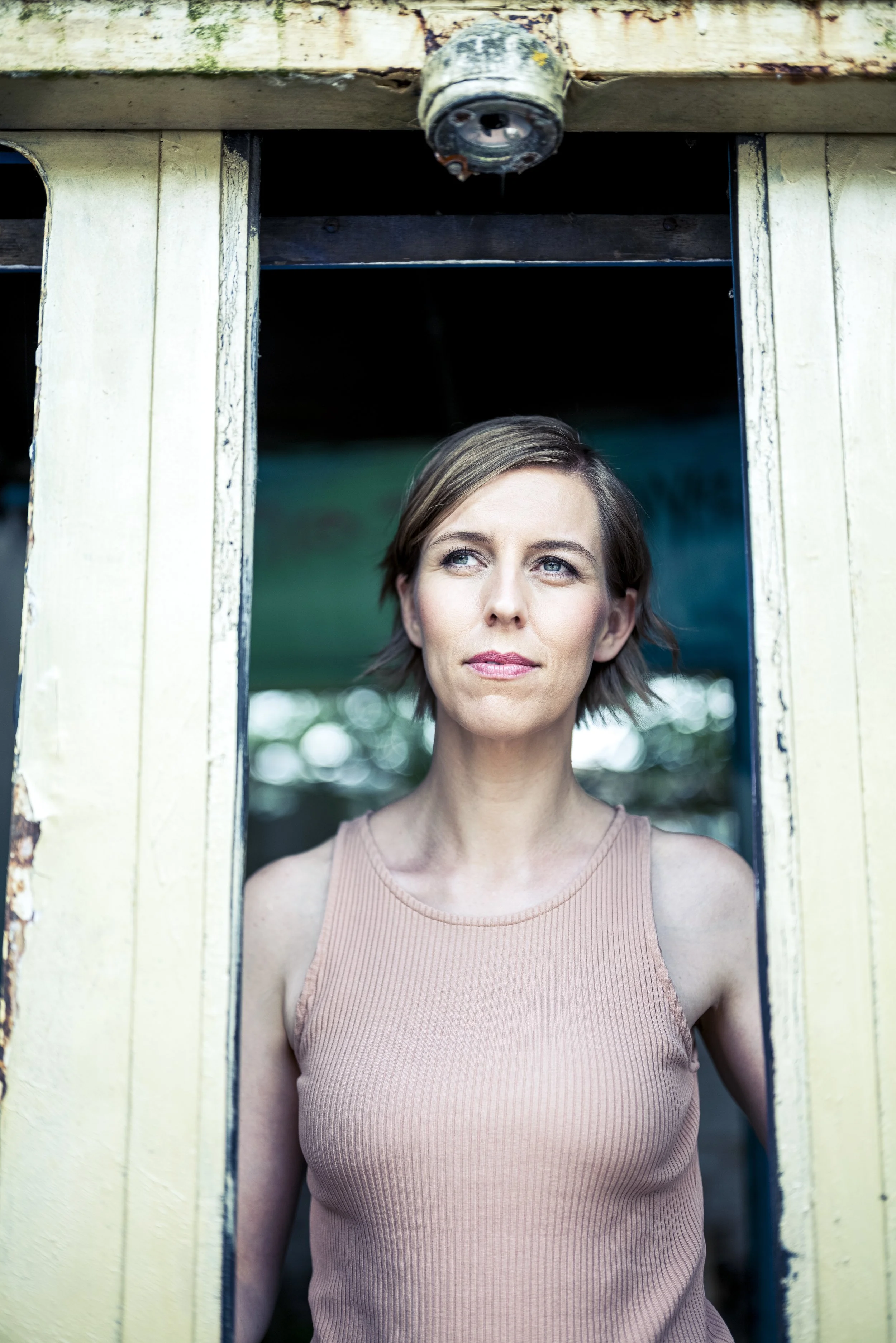 Eine Frau mit kurzen braunen Haaren steht in einem alten, verfallenen Gebäude, Blick nach oben, umgeben von einem grünen Hintergrund.