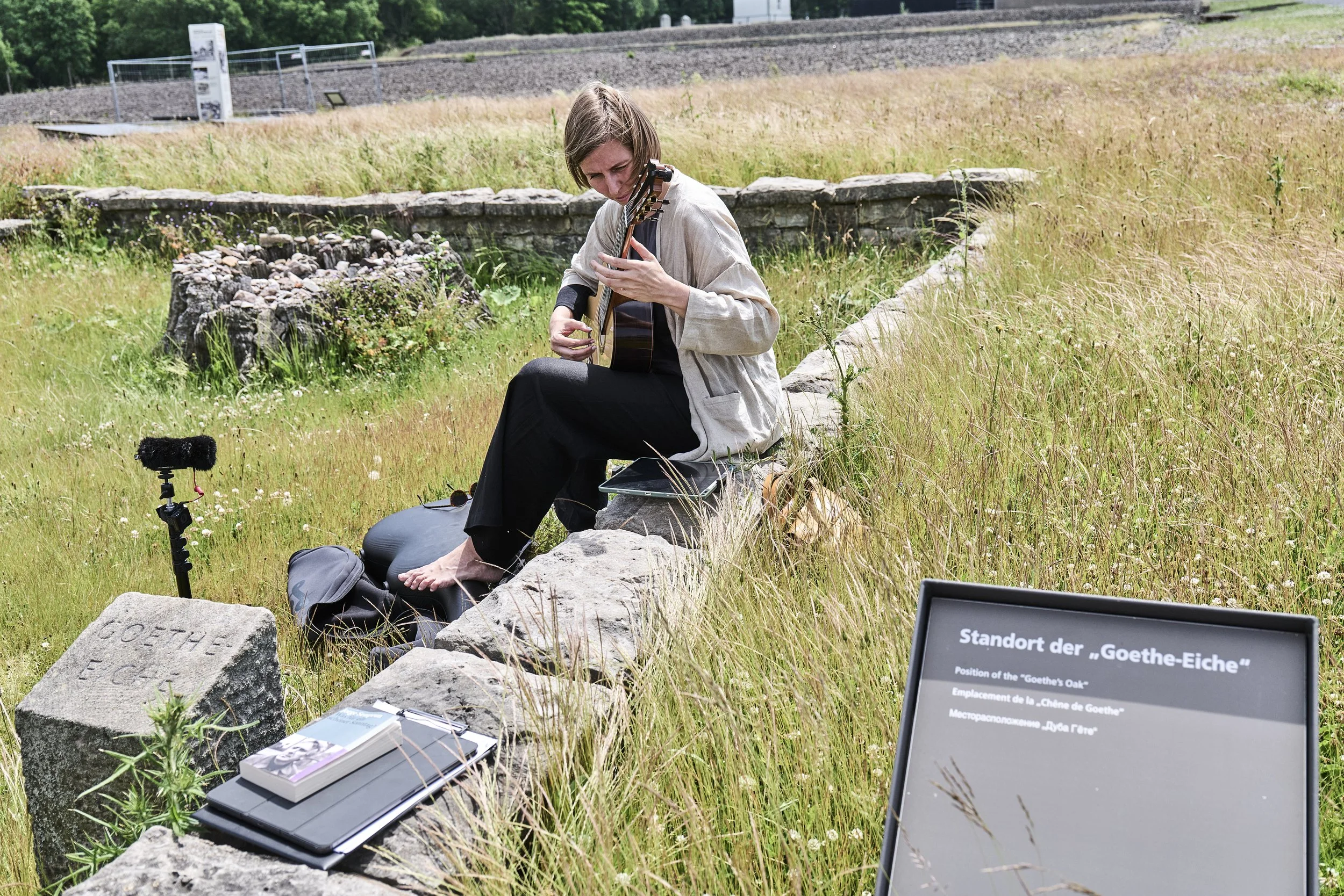 Frau spielt Gitarre im Freien, umgeben von Gras und Steinen, mit Aufnahmegeräten und einem Informationsschild über den Standort der Goethe-Eiche.