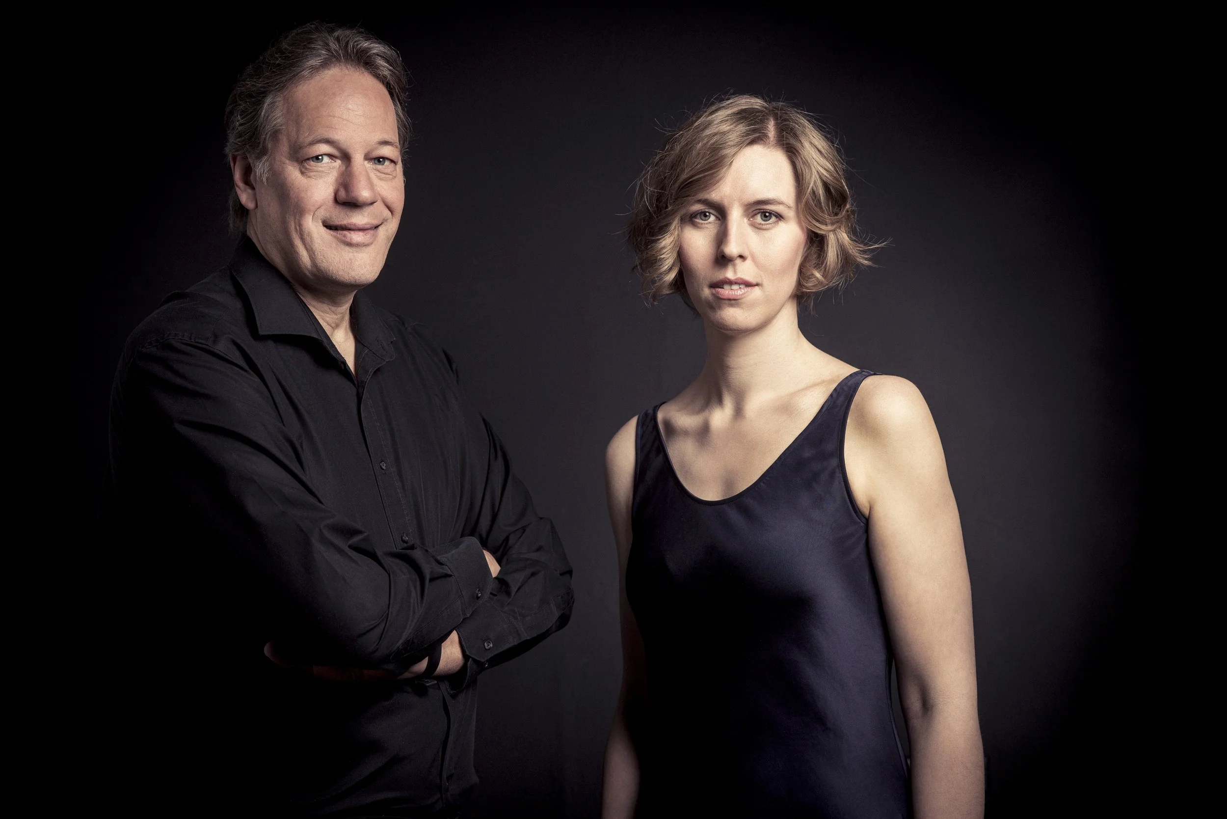 Ein Mann in schwarzer Hemd und eine junge Frau in einem schwarzen Kleid stehen nebeneinander vor einem dunklen Hintergrund.