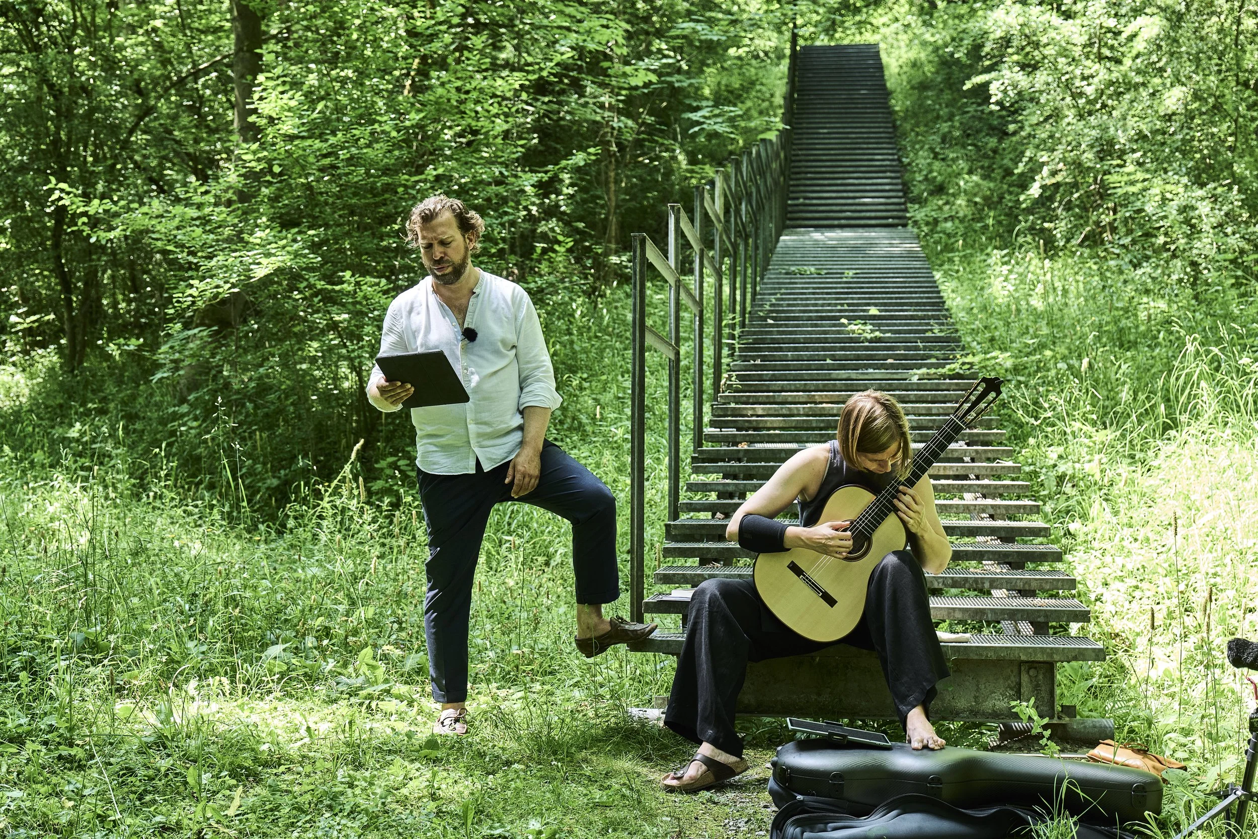 Zwei Musiker im Grünen, einer steht mit Tablet, die andere spielt Gitarre auf einer Bank, im Hintergrund eine Steintreppe.