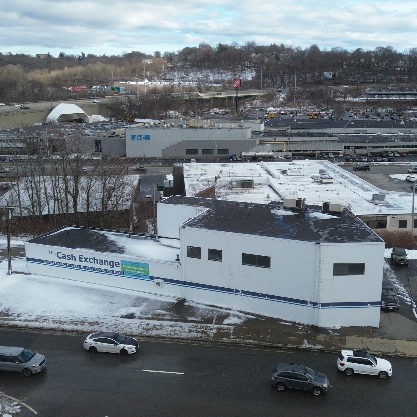 Cityscape view showing a building with a sign that reads 'The Cash Exchange' and a few cars on the street in front, snow on rooftops, and a commercial area with parking lots and factory buildings in the background.
