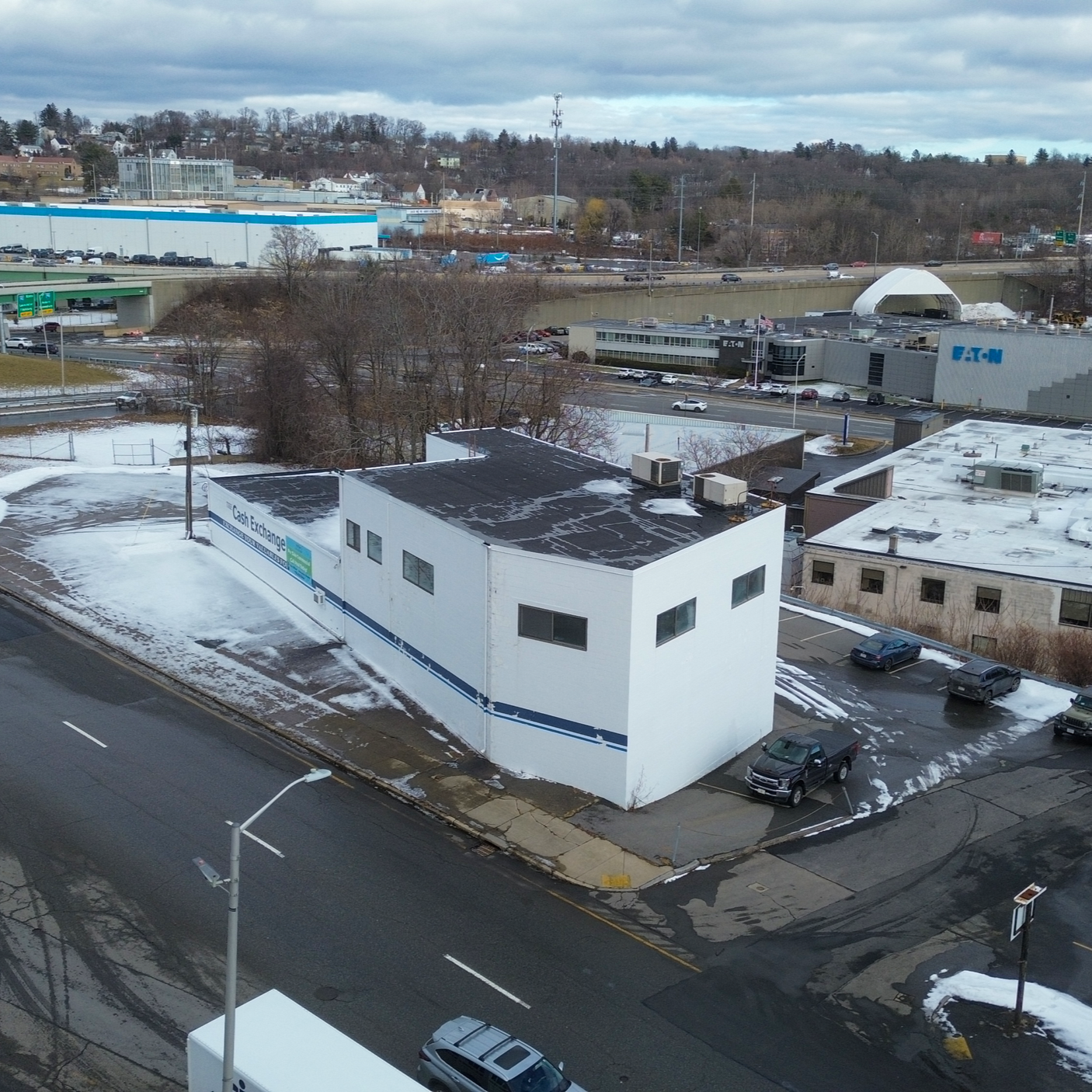 A white commercial building with a sign reading 'Cash Exchange' on the side, parked with some snow and ice on the ground in an urban area. Surrounding roads have some parked cars and light snow patches.
