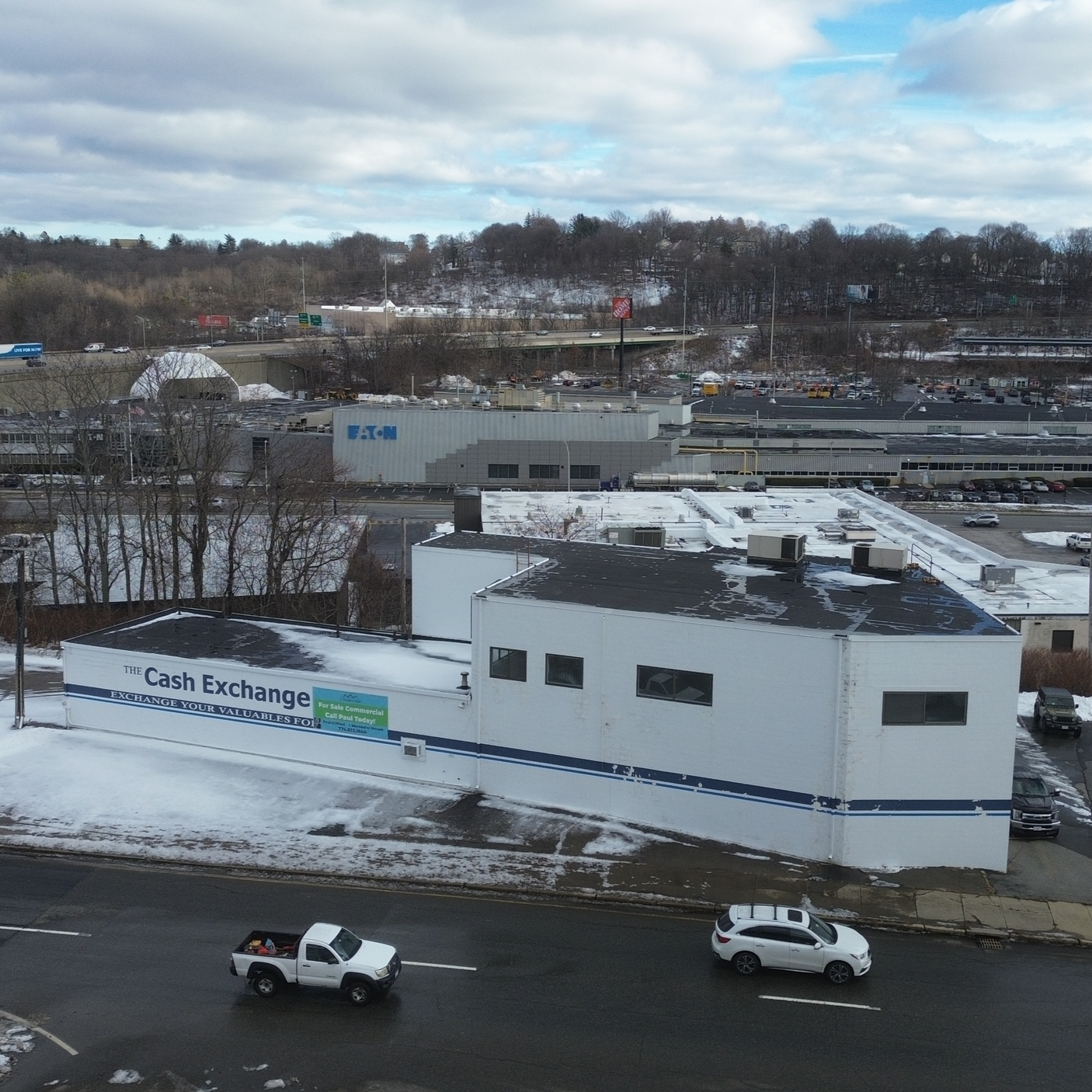 A snow-covered commercial building labeled 'The Cash Exchange' on a city street with two cars parked and a few vehicles driving by. In the background, a shopping area with the 'EAT' store visible, and beyond, a parking lot and hilly, partly snow-cove