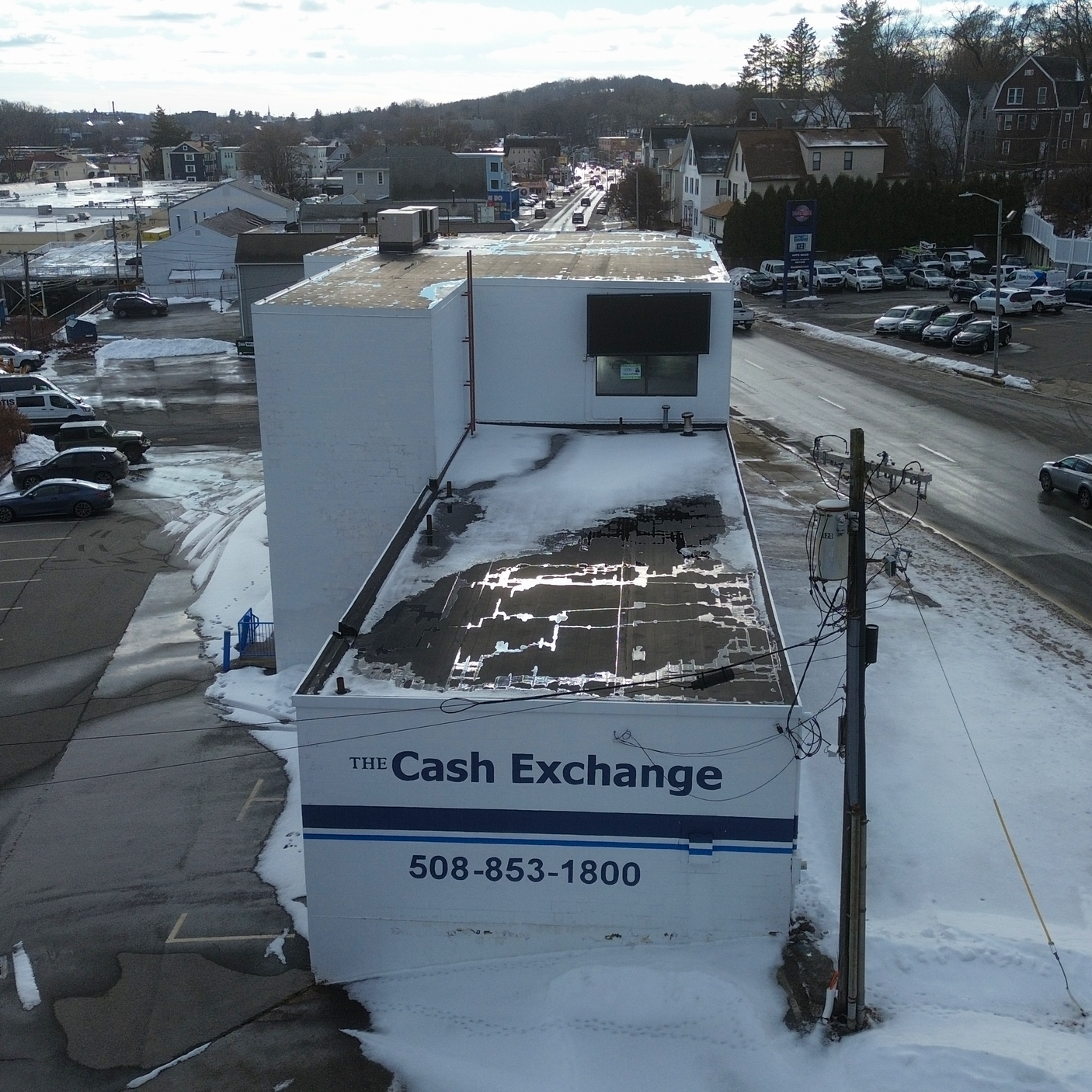 A rooftop view of a building with a sign that reads 'The Cash Exchange' and a phone number, with snow on the roof and parking lot, and a street with cars and houses in the background.
